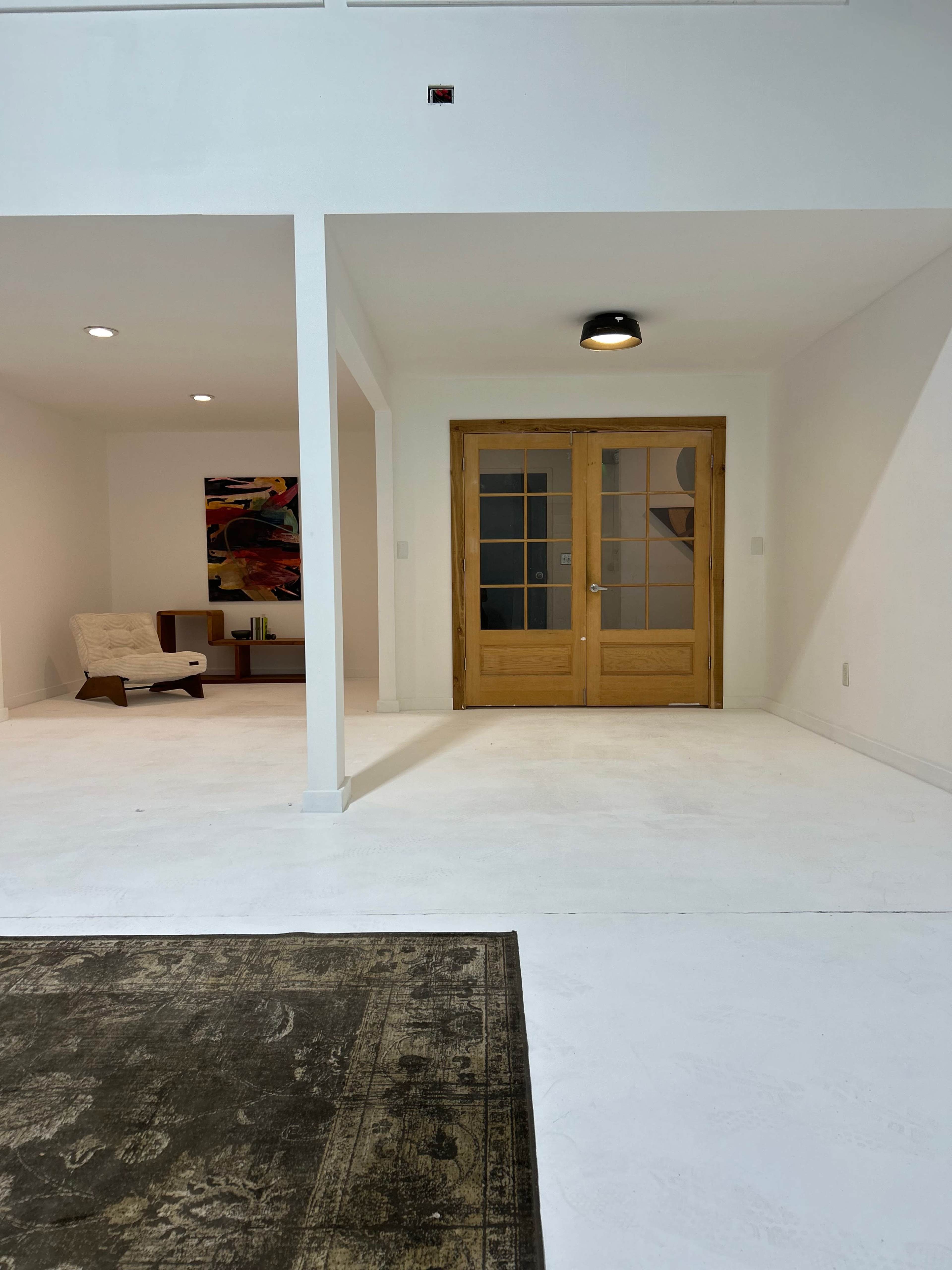 The image shows a spacious, modern interior with a doorway leading to another room, a light fixture overhead, and a patterned rug on the white floor.