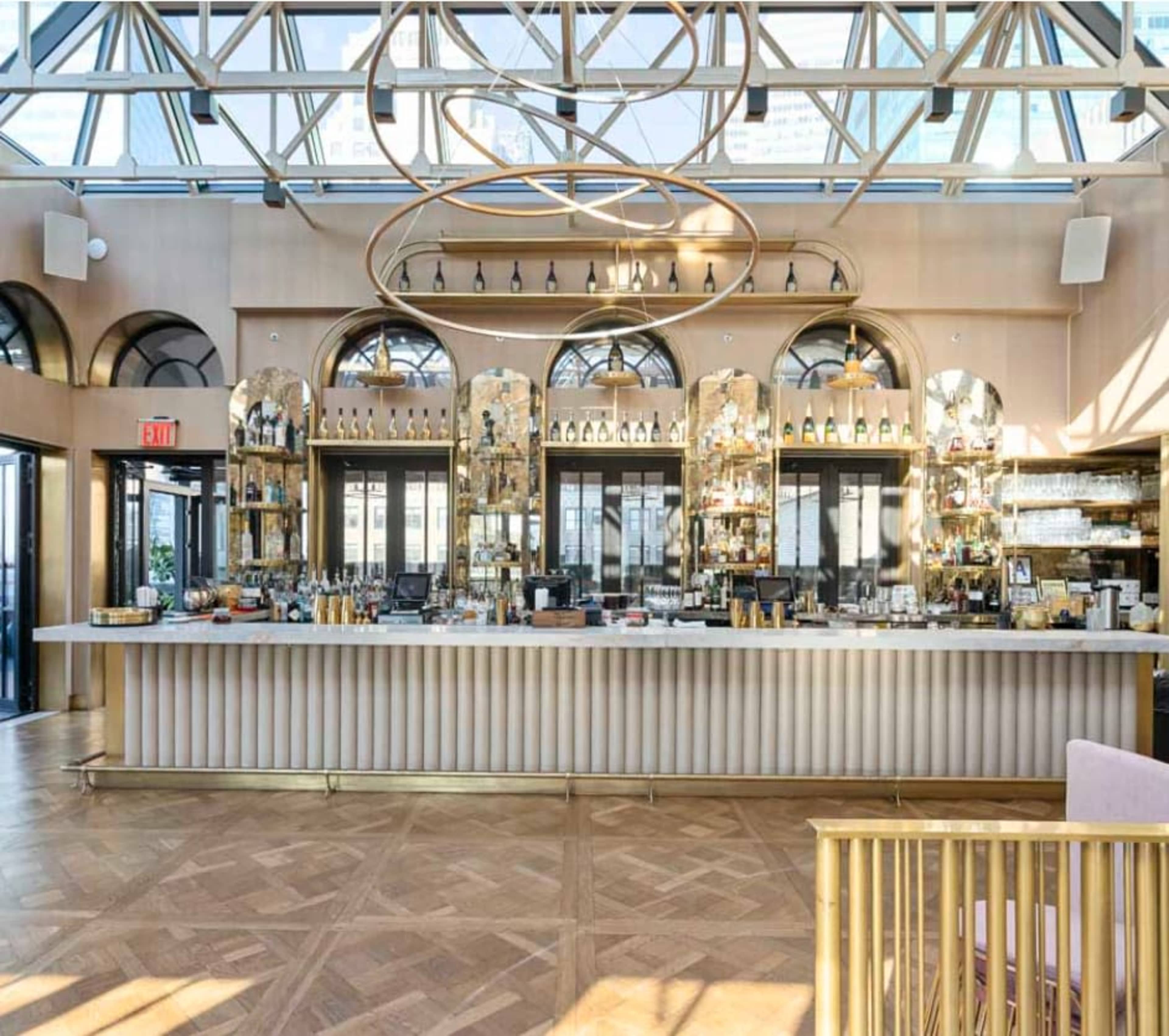 The image shows a modern bar area with a long countertop, a selection of bottles displayed on shelves, and large windows overhead allowing natural light to illuminate the space.