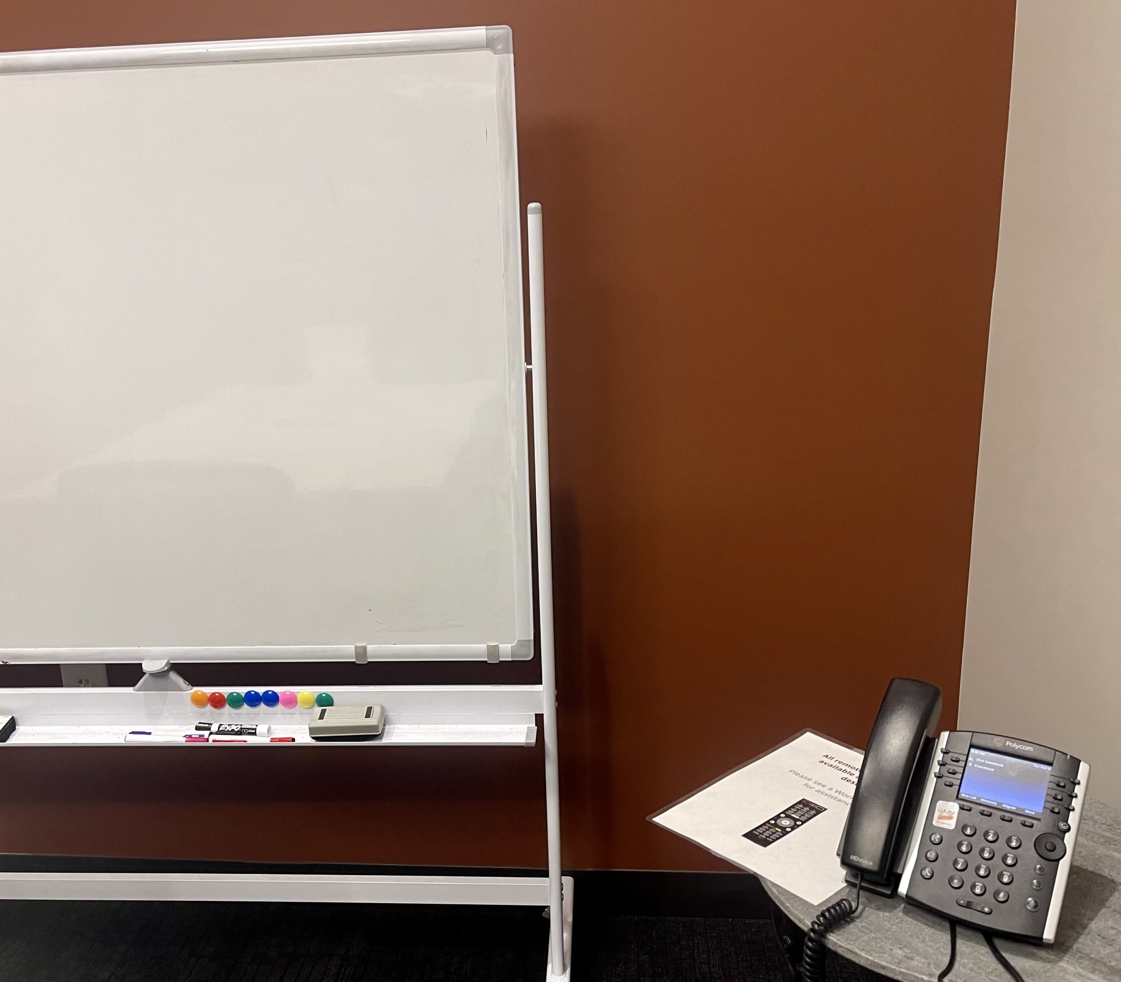 A whiteboard on a stand next to a desk with a telephone and a piece of paper.