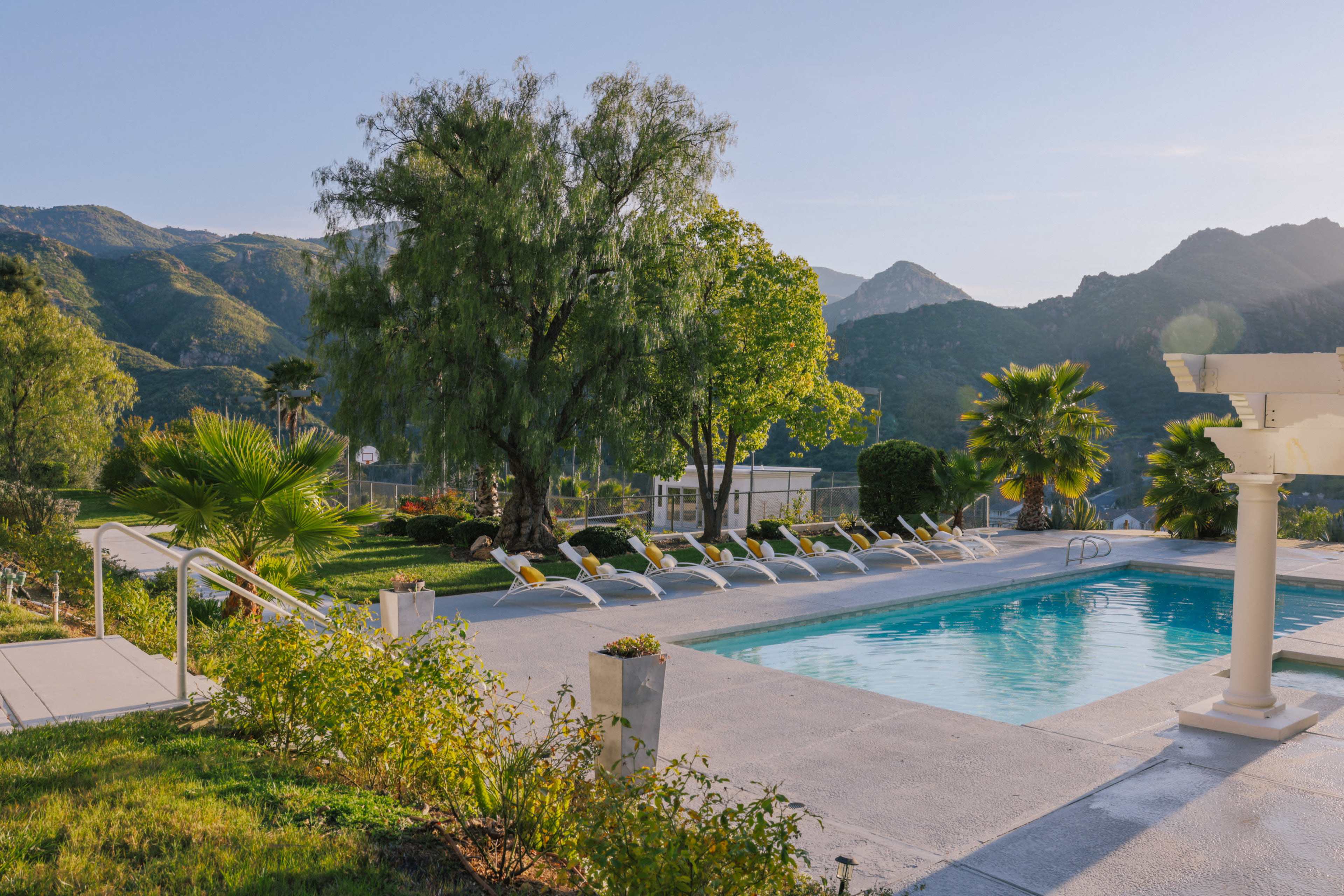 A swimming pool surrounded by neatly arranged lounge chairs, with mountains and greenery in the background.
