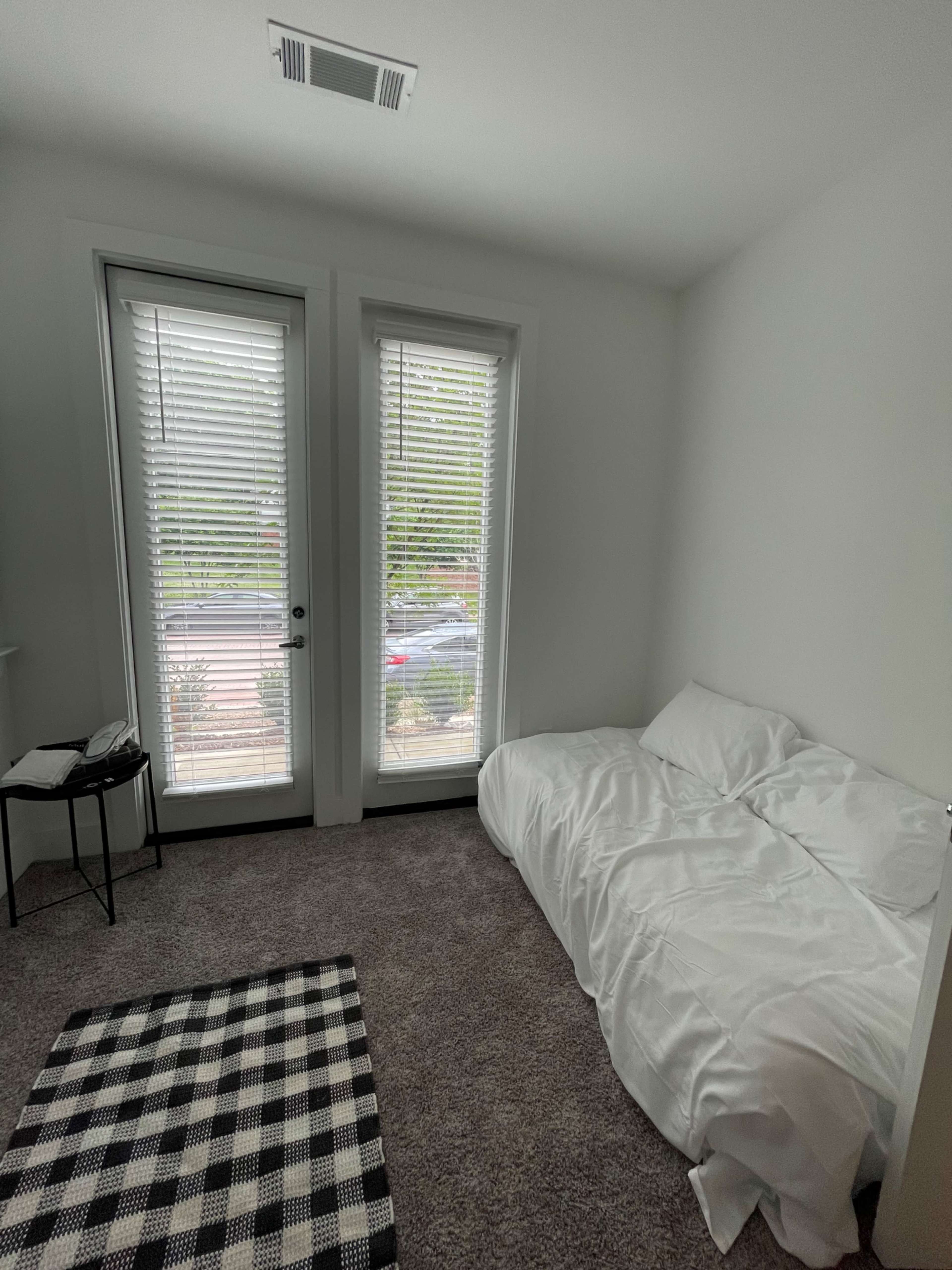 A simple bedroom features a bed with white bedding, a checkered rug on the carpeted floor, and two windows with blinds.