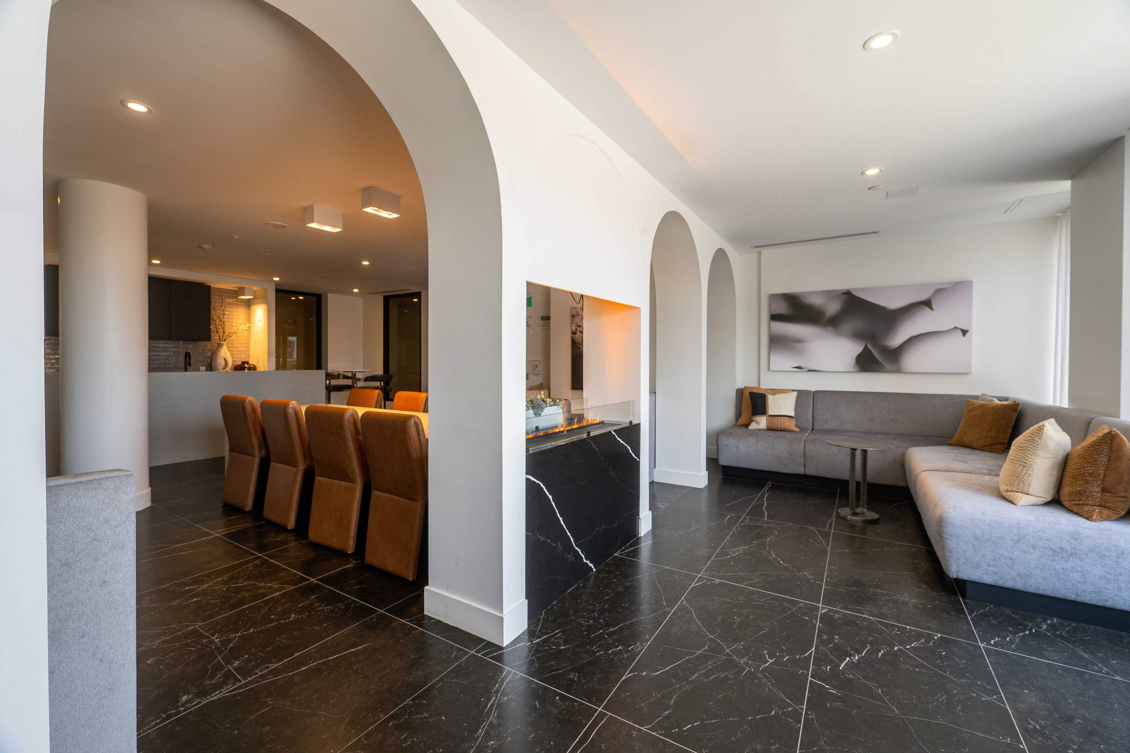 The image shows a modern lounge area featuring a sectional gray couch, a round table, and a sleek black marble fireplace, with a dining area that includes brown upholstered chairs in the background.
