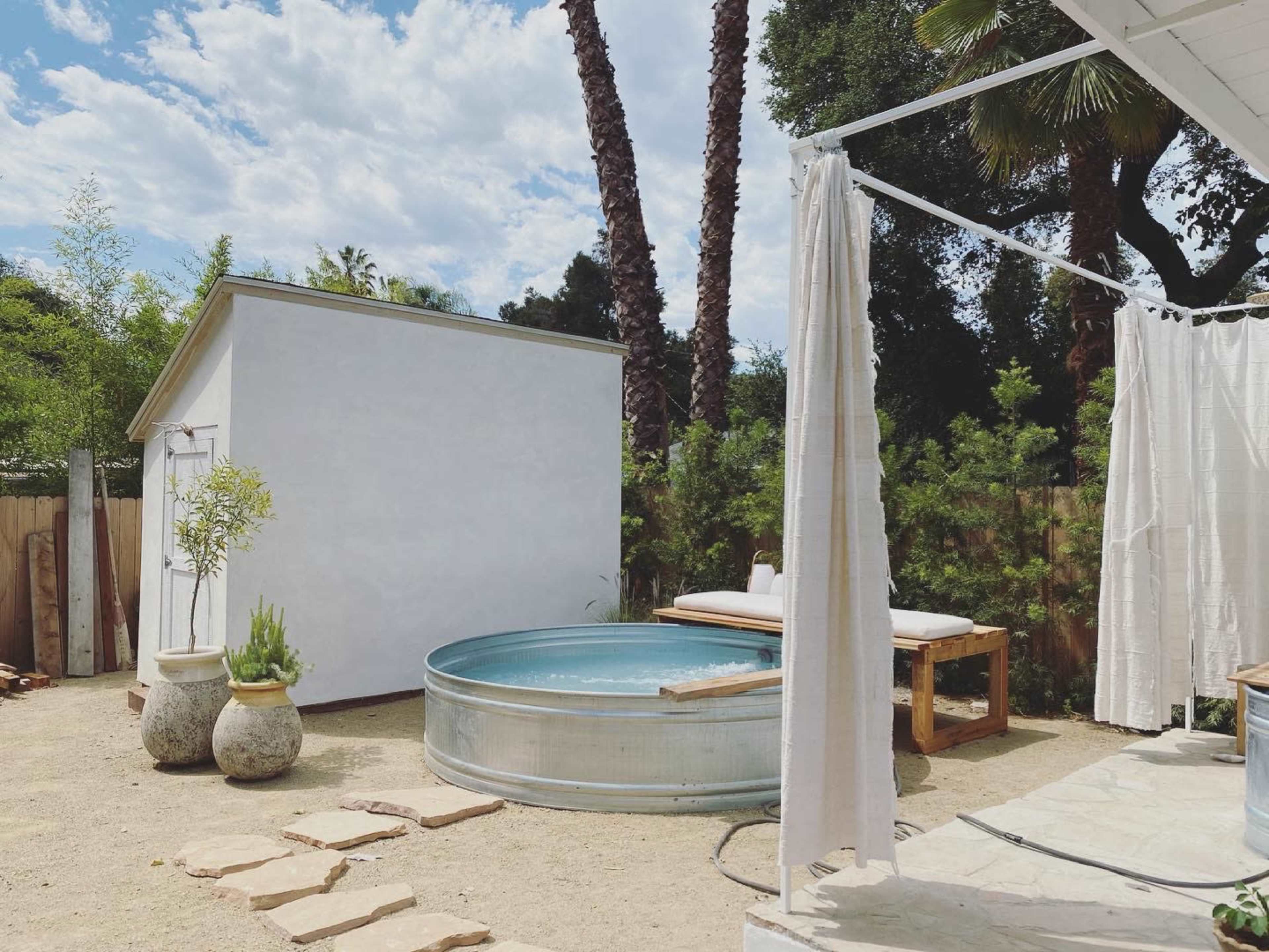 A round metal tub is situated on a sandy patio surrounded by palms and a white shed, with light-colored curtains draping nearby.