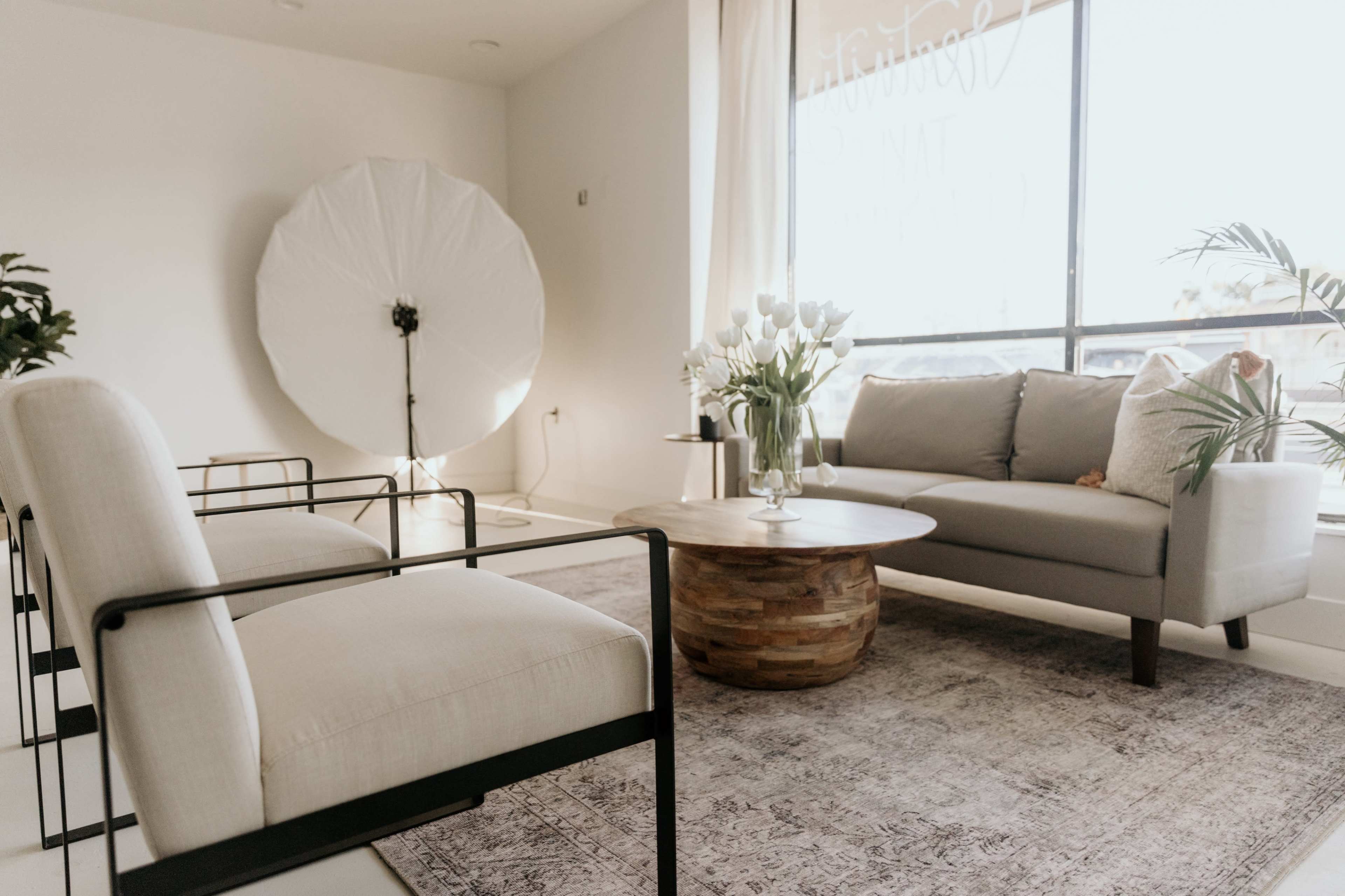 A modern, minimalistic living room with a gray sofa, two light-colored chairs, a round wooden coffee table, and a large light reflector in the background.
