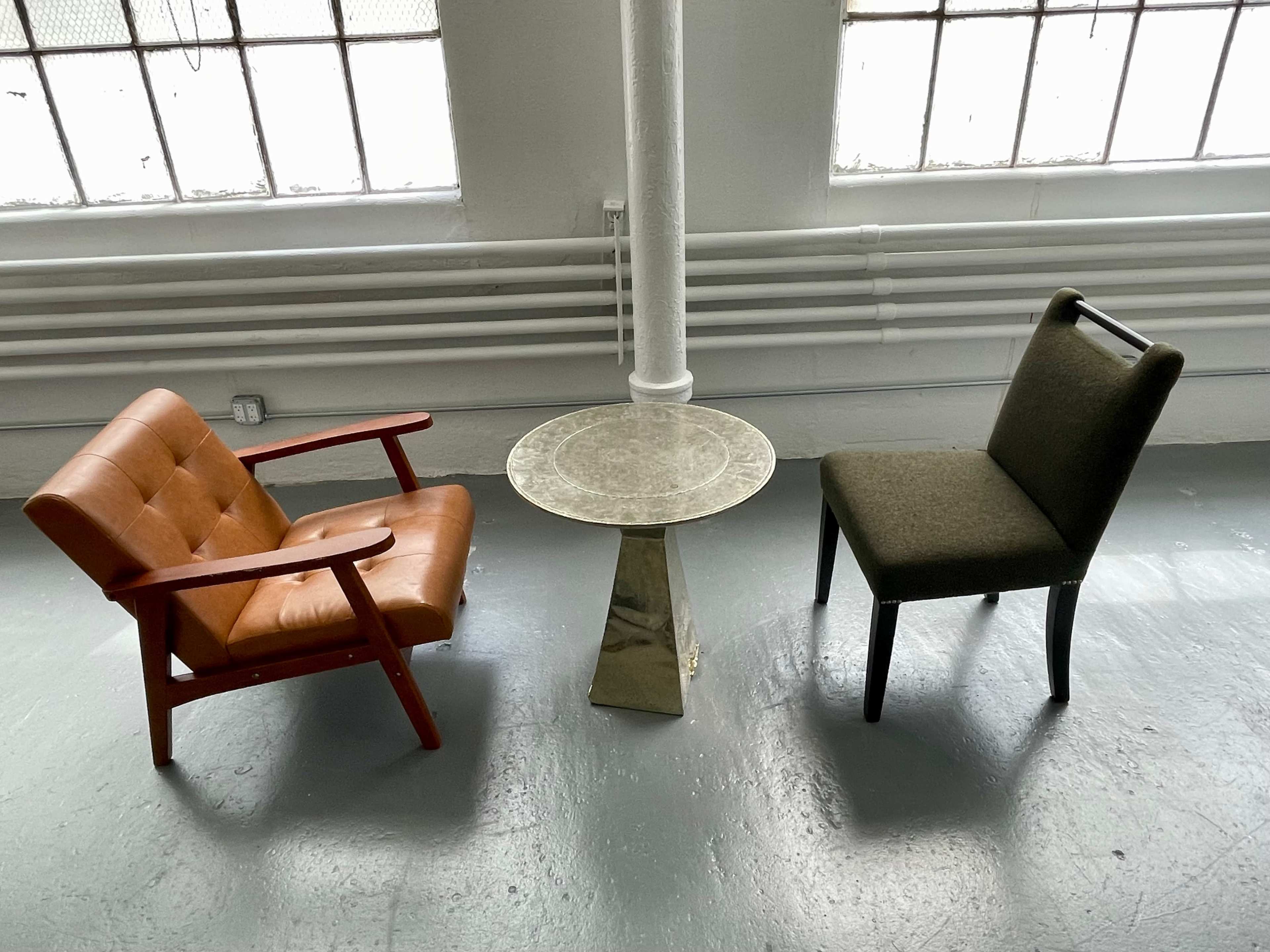 The image shows two chairs—a brown armchair and a green upholstered chair—separated by a round, metallic side table in a room with large windows.