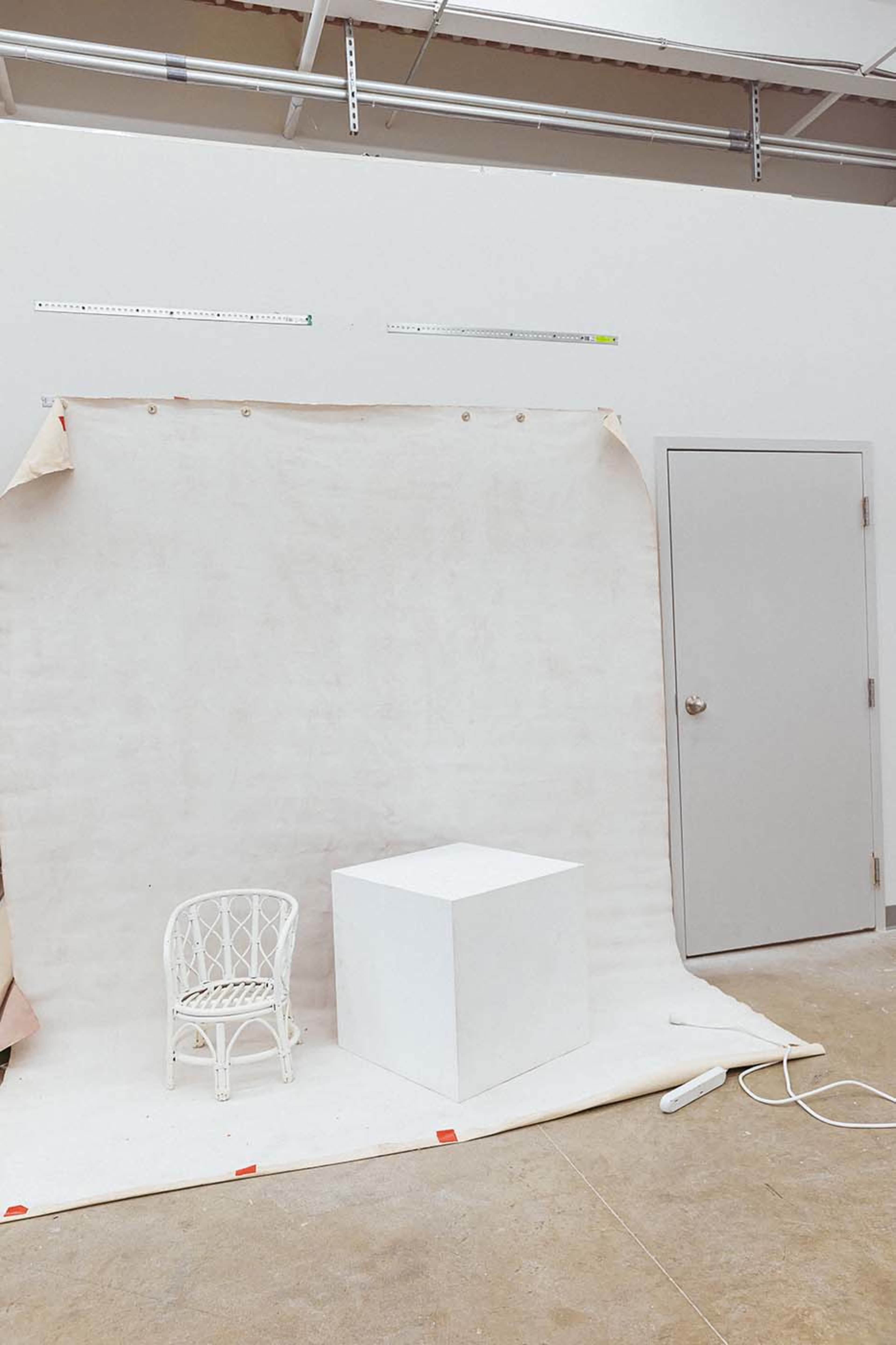 A white cube and a small white chair are positioned in front of a plain, light-colored backdrop in a studio space.