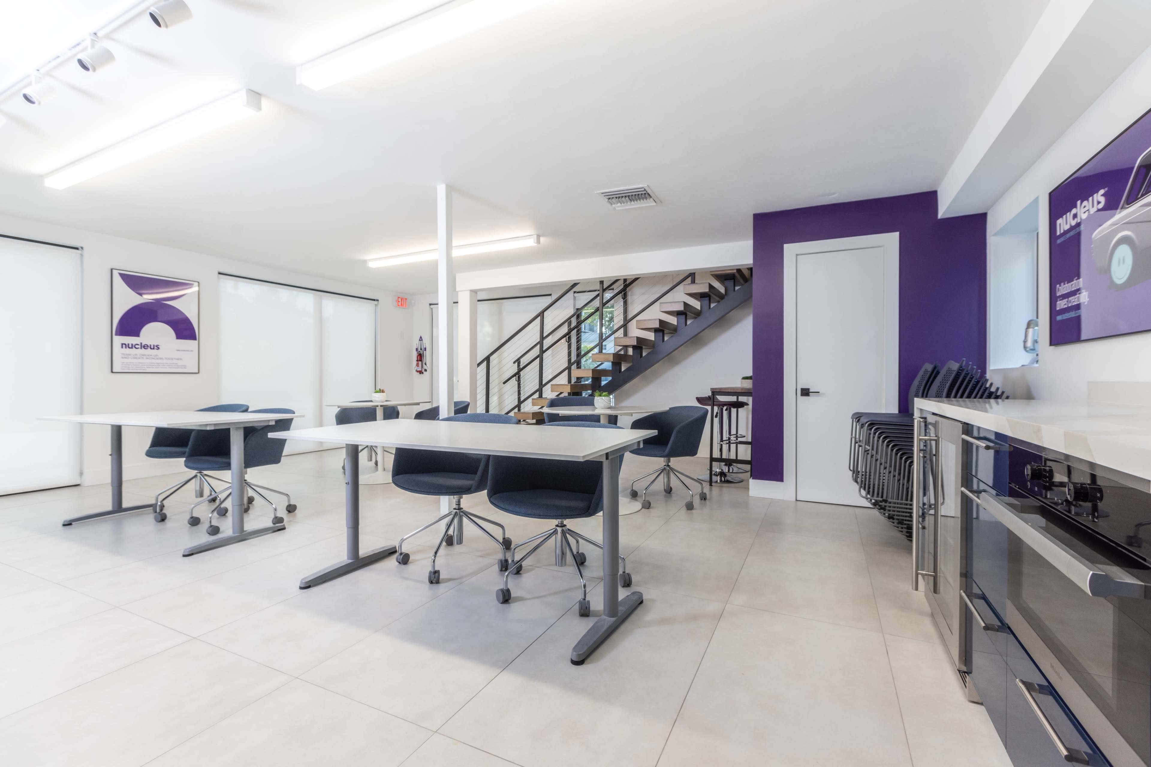 A modern, brightly lit room with several tables and chairs set up for a meeting, along with a staircase and kitchen area in the background.