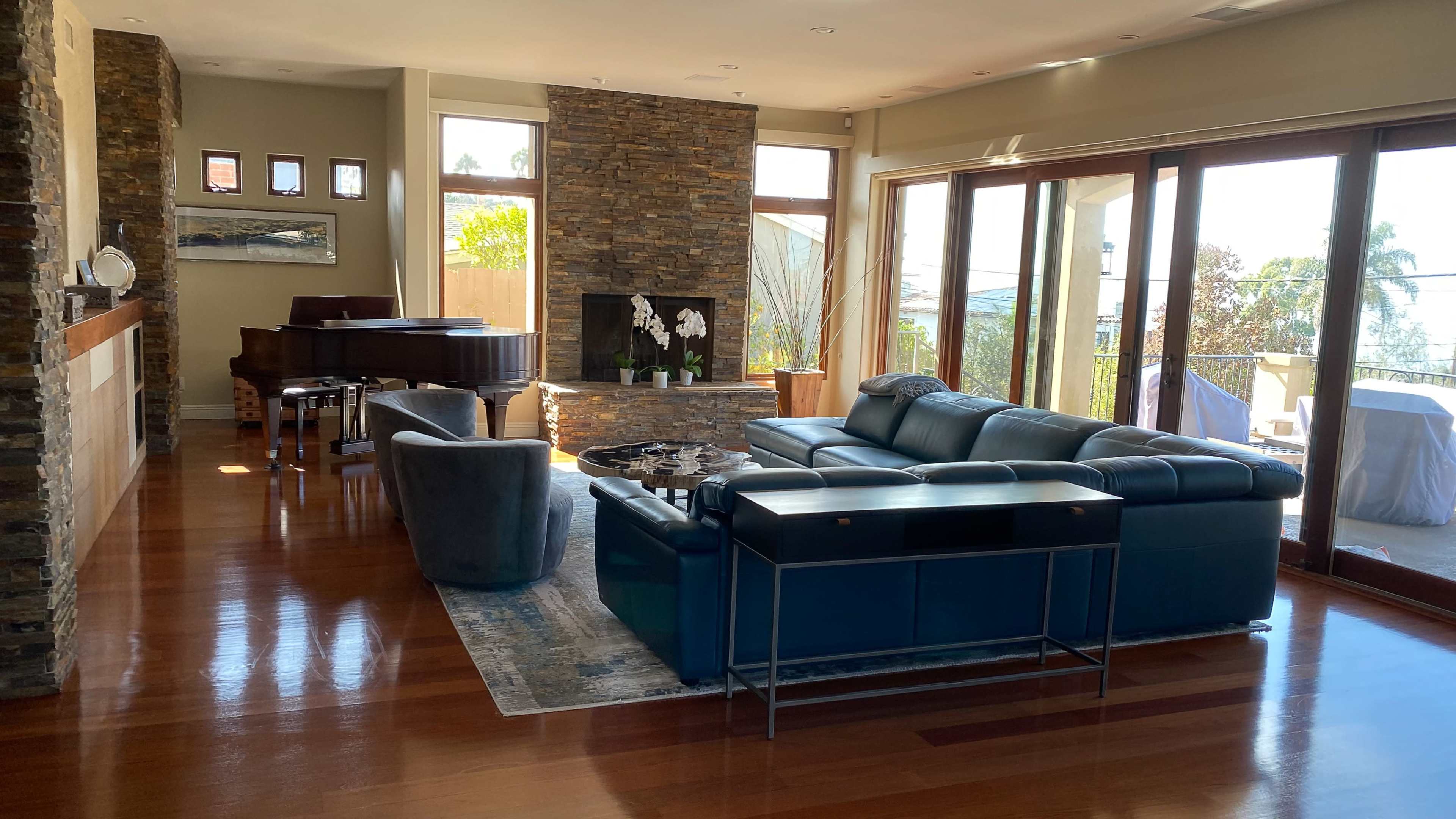 The image shows a spacious living room featuring a stone accent wall, a grand piano, a large sectional sofa, and floor-to-ceiling windows that let in ample natural light.