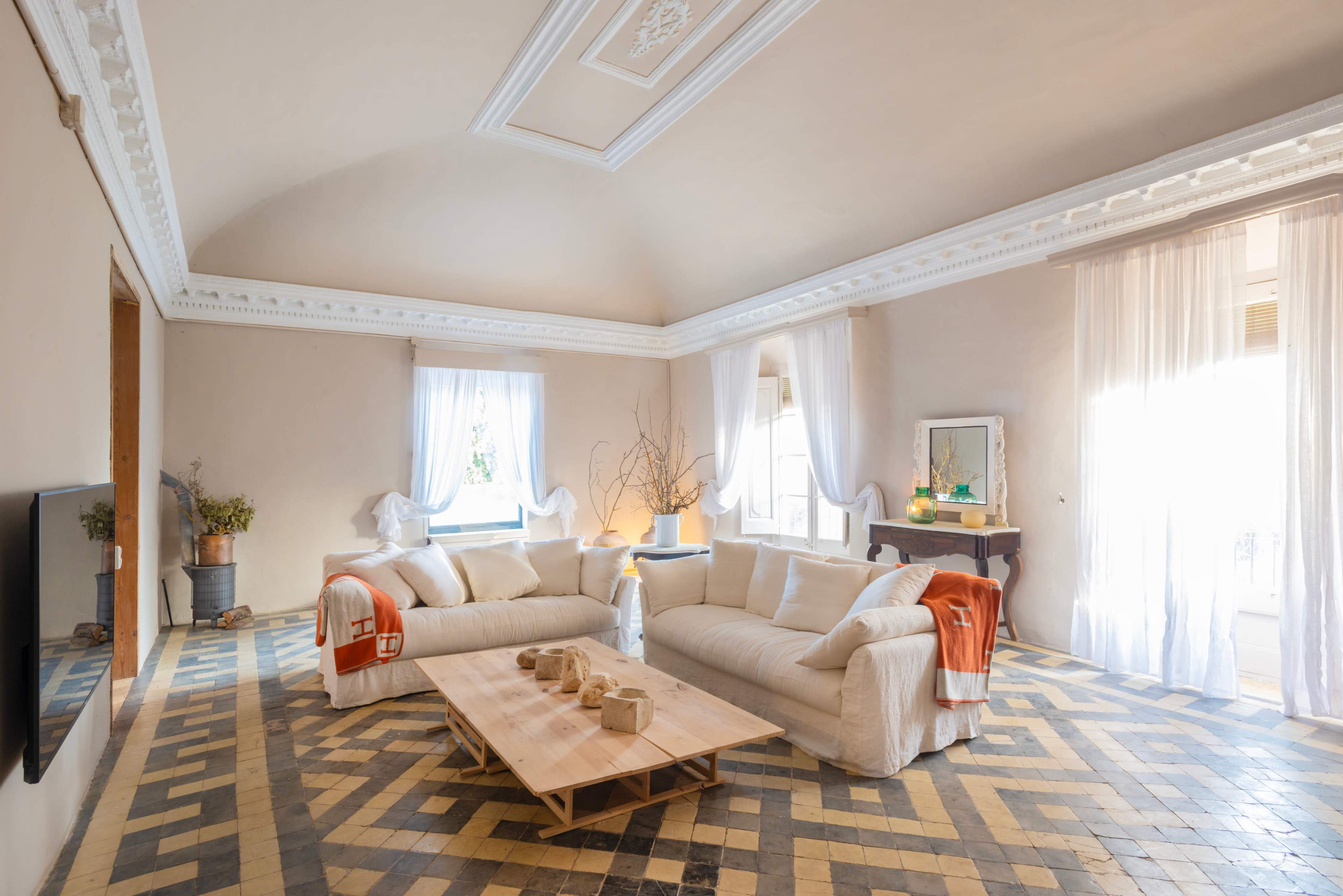 The image shows a spacious living room with a light-colored ceiling, two white sofas, a wooden coffee table, and large windows allowing natural light to enter.