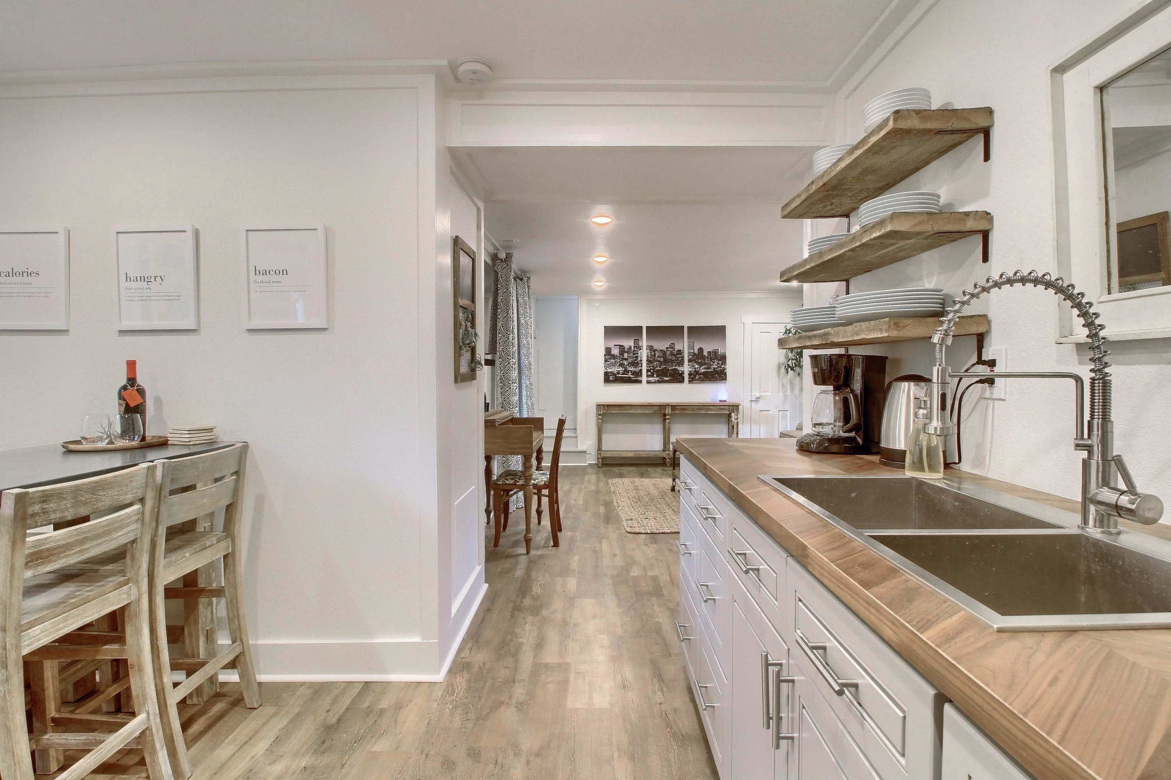 A modern kitchen features a wooden counter with bar stools, open shelving with dishes, and a view into a dining area with a table and artwork on the walls.