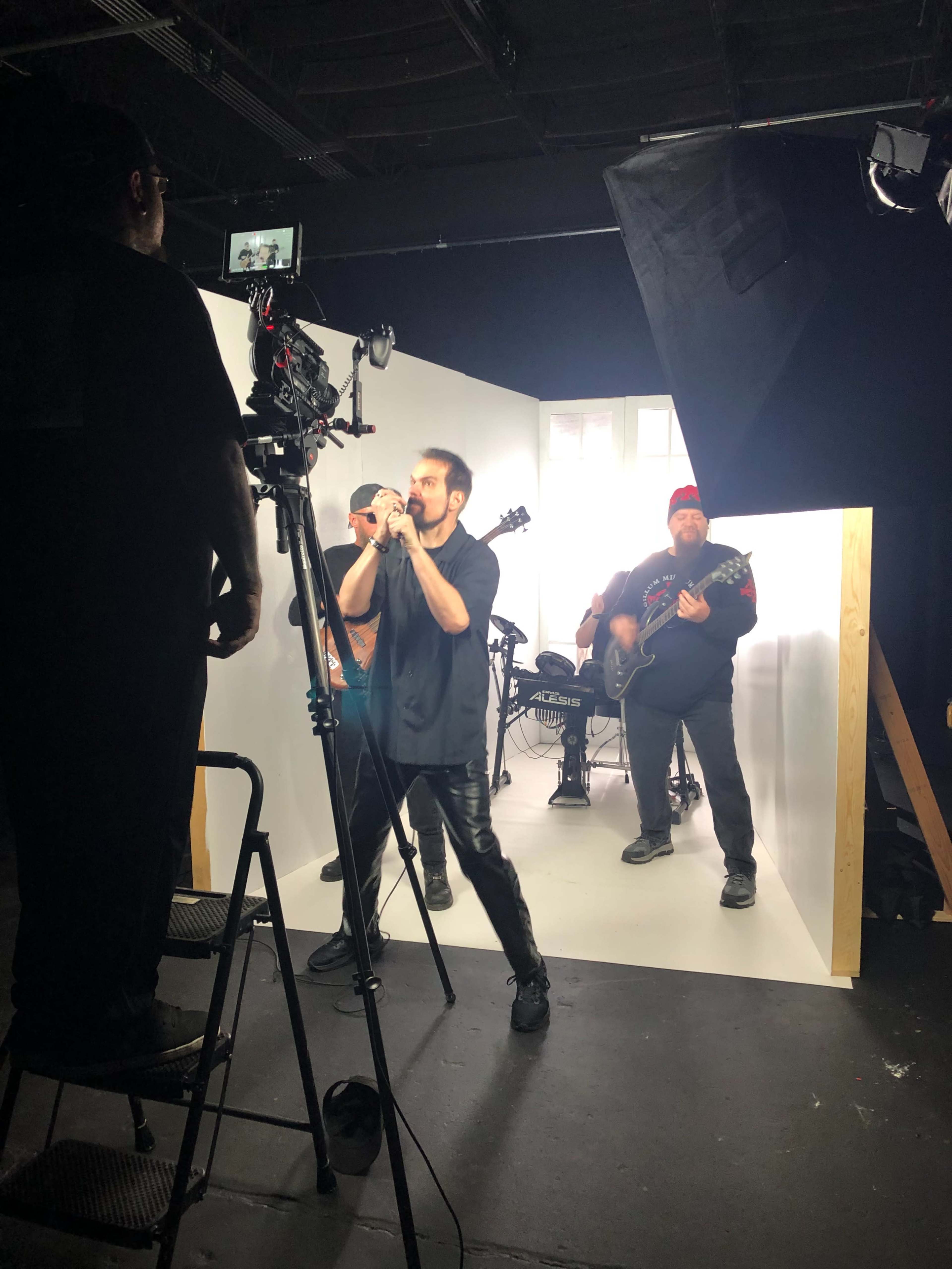 A musician performs in front of a camera while a band plays in the background on a stage with white backdrops.