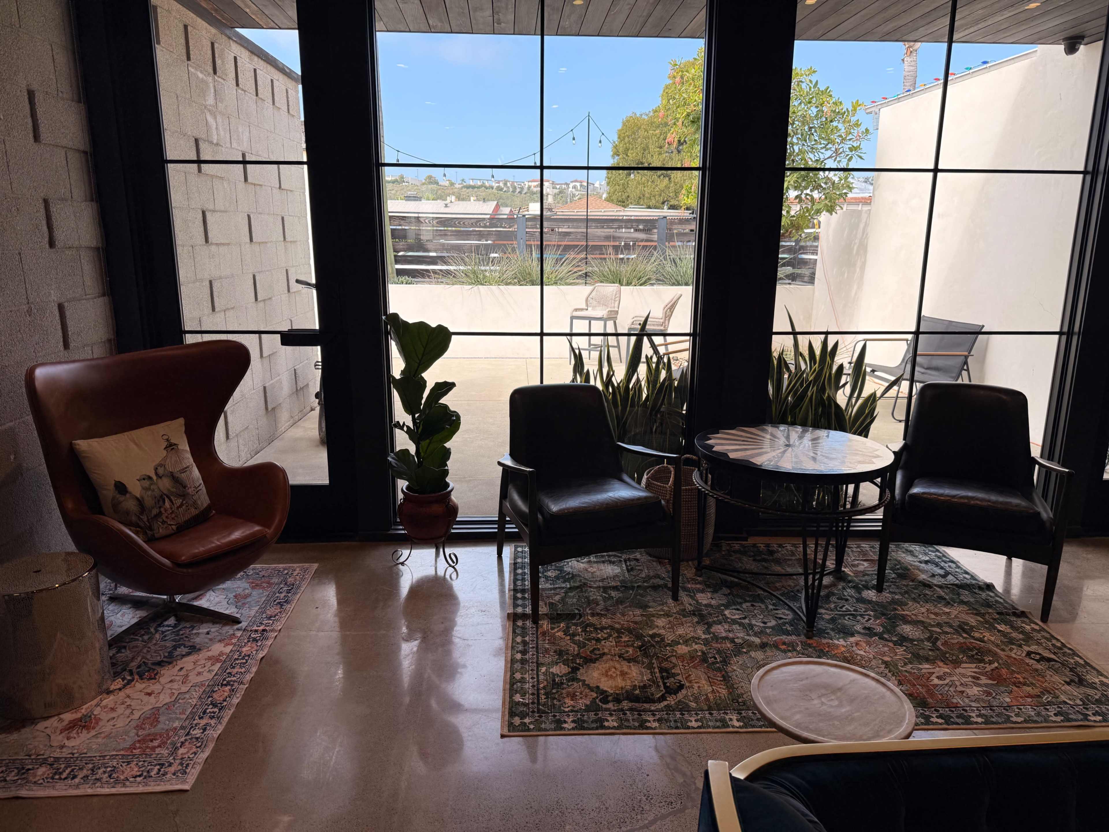 A cozy indoor seating area features two chairs, a round table, a potted plant, and large windows overlooking an outdoor space with greenery.