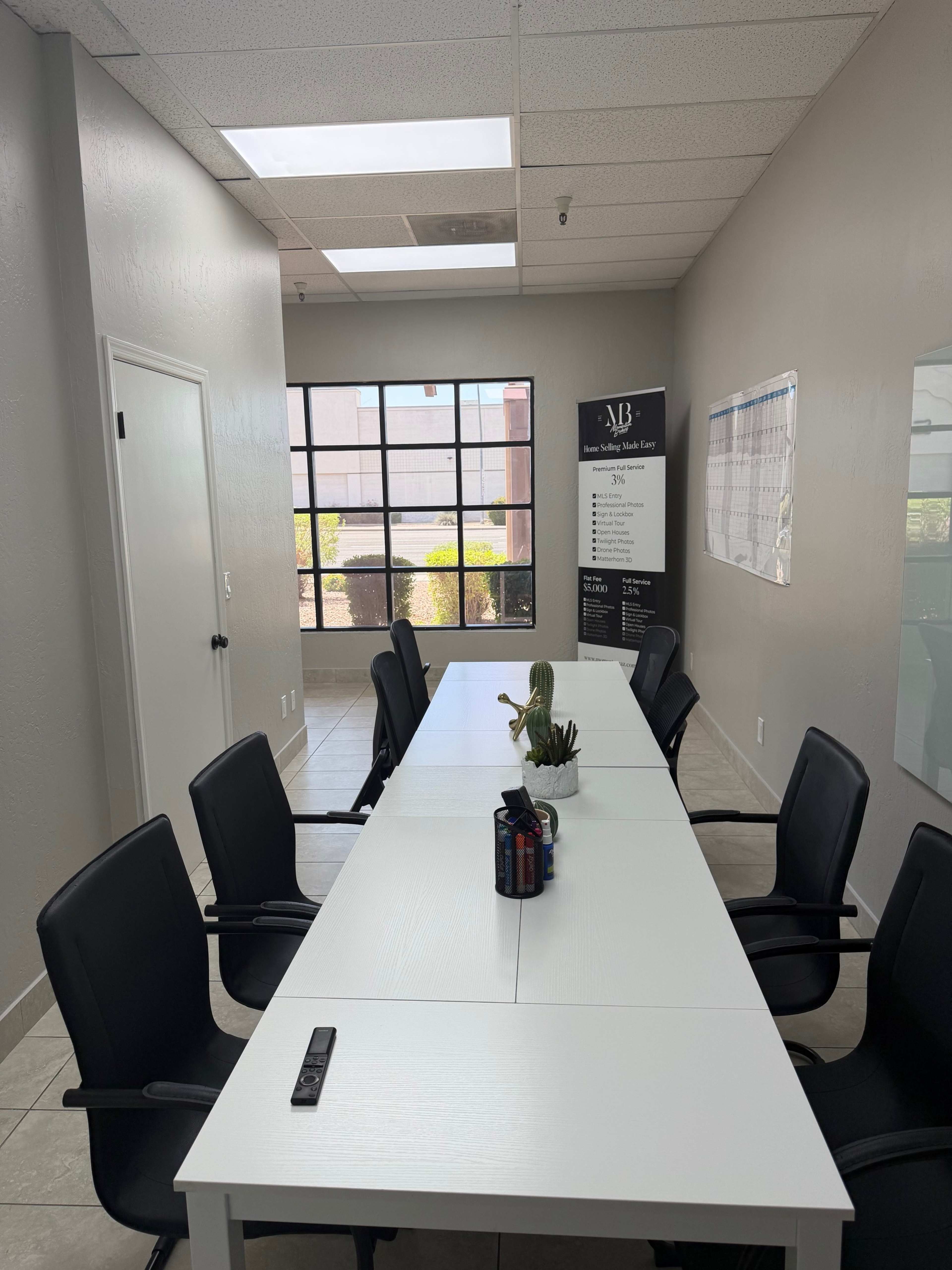 The image shows a modern conference room with a long white table surrounded by black chairs, large windows letting in natural light, and a banner on one side.