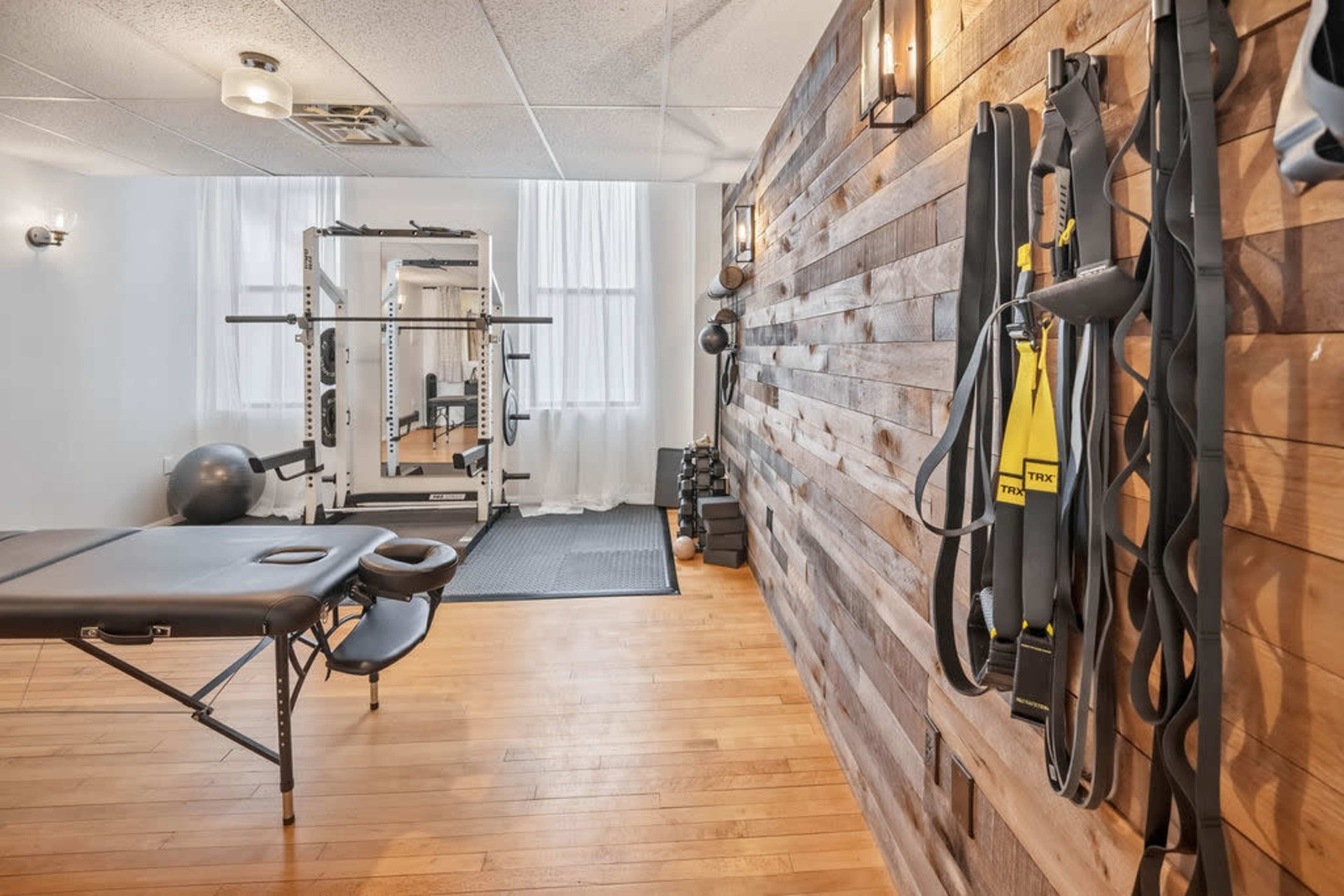 The image shows a home gym with wooden walls, exercise equipment, a therapy table, and various fitness accessories.