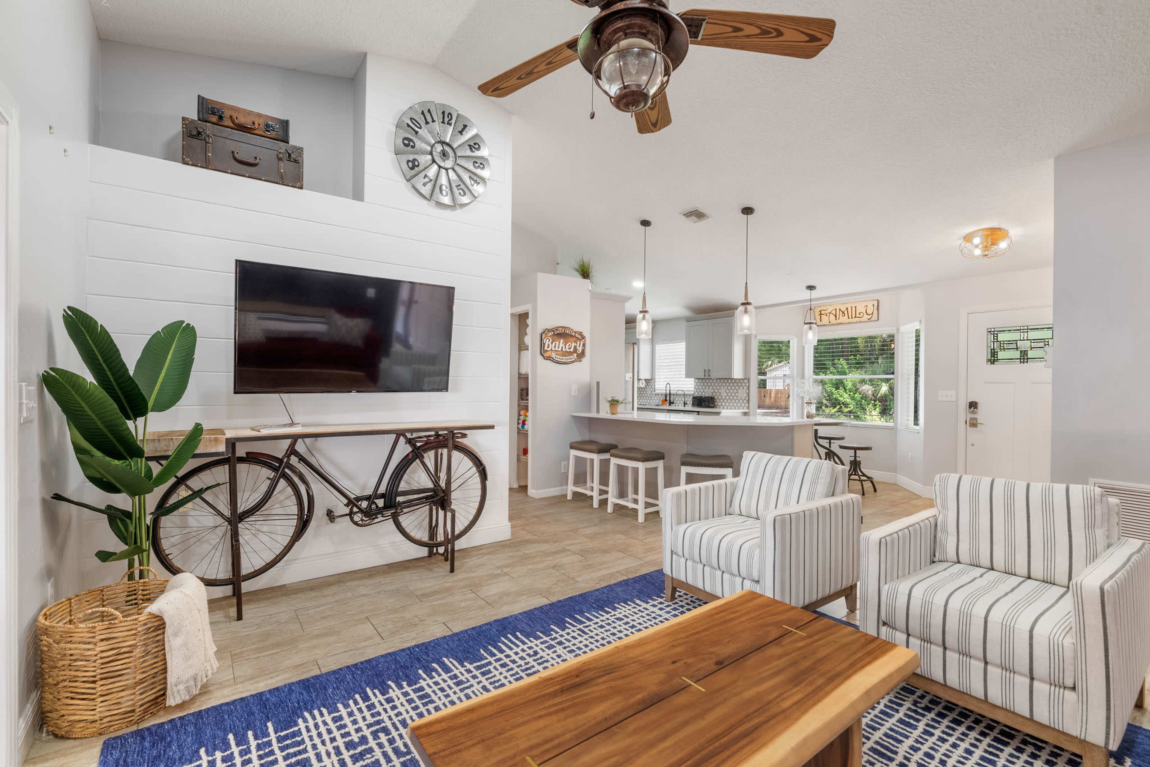 A modern living space featuring a TV mounted on a white shiplap wall, a decorative bicycle, two striped chairs, and an open kitchen area.