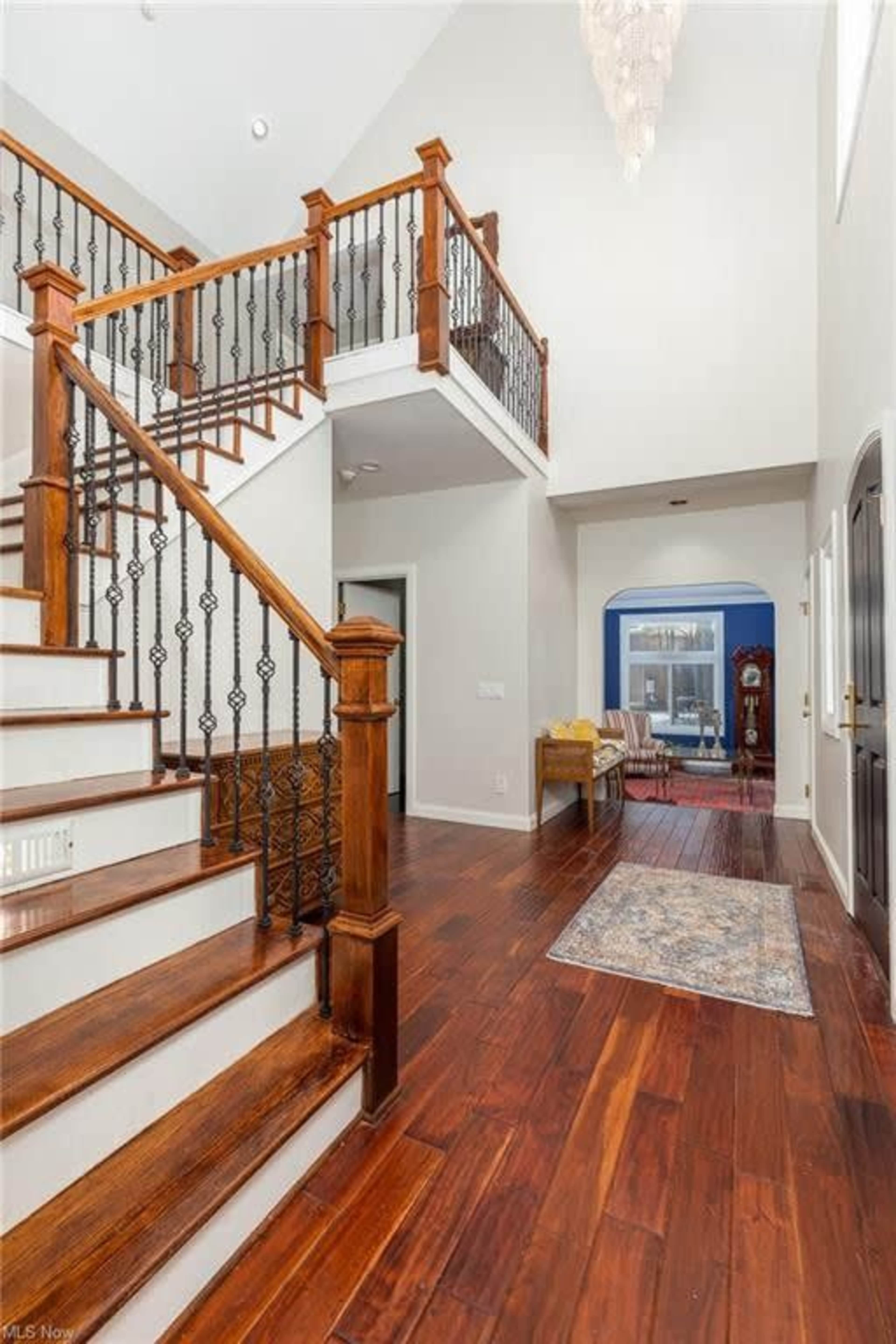 A spacious foyer with a wooden staircase, decorative railing, and hardwood flooring leading to an arched doorway in the background.