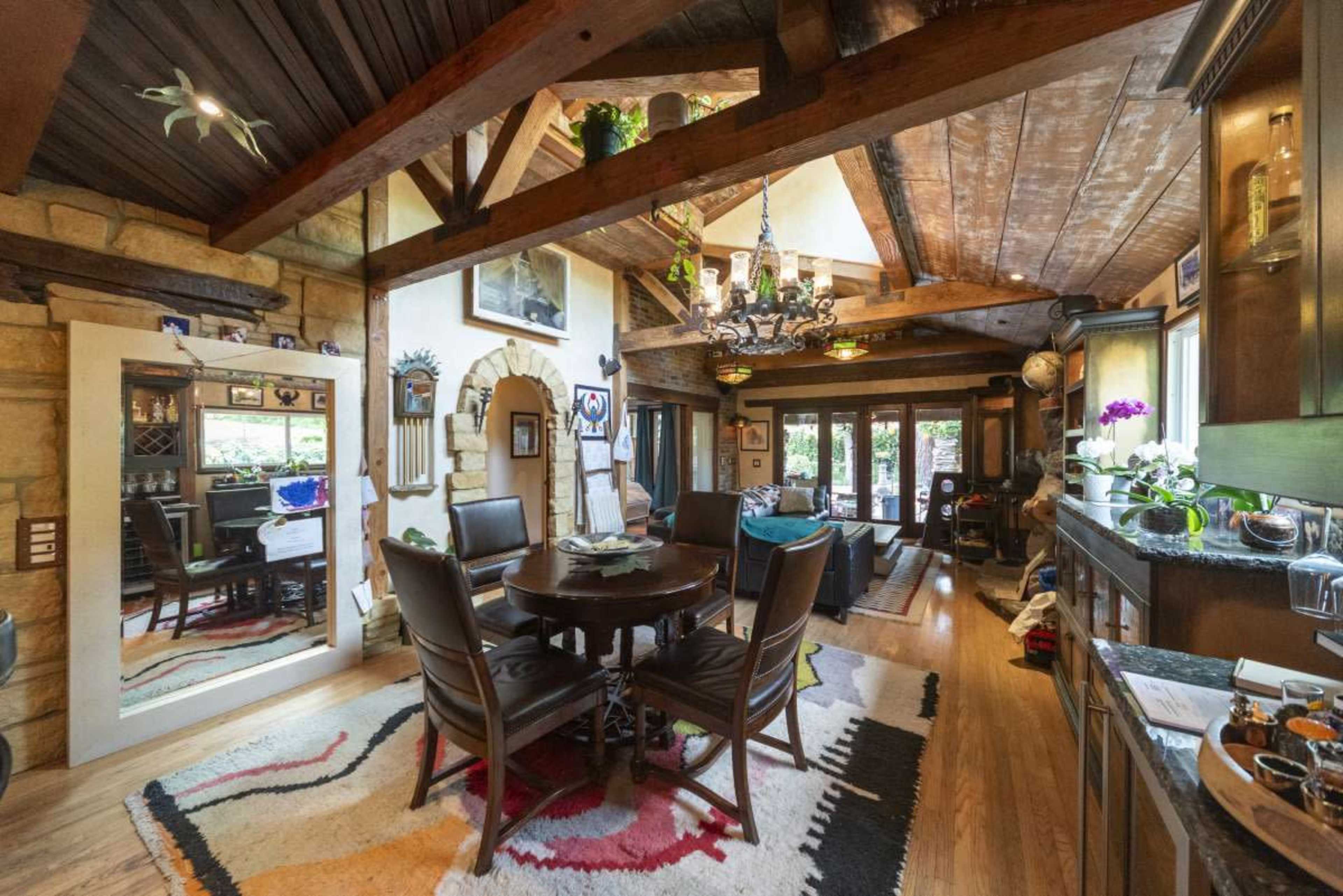 The image shows a rustic dining area with a wooden table surrounded by chairs, adorned with various decorative elements and a stone wall.