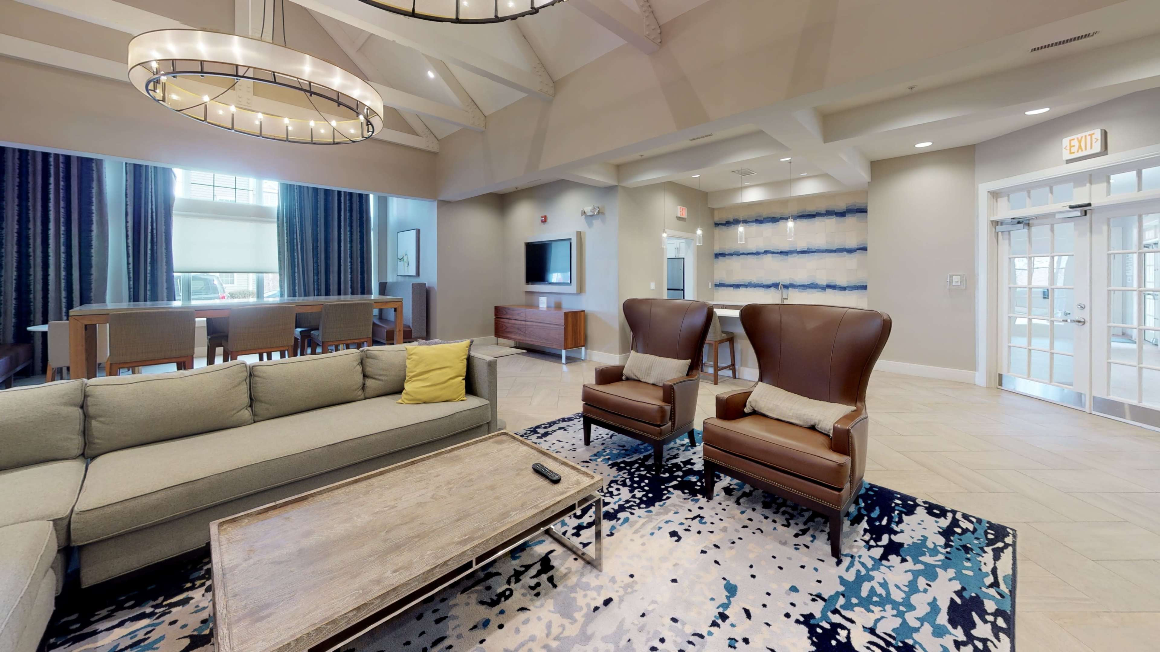 A modern lounge area featuring a light gray sofa, two brown armchairs, a wooden coffee table, and a television on the wall.