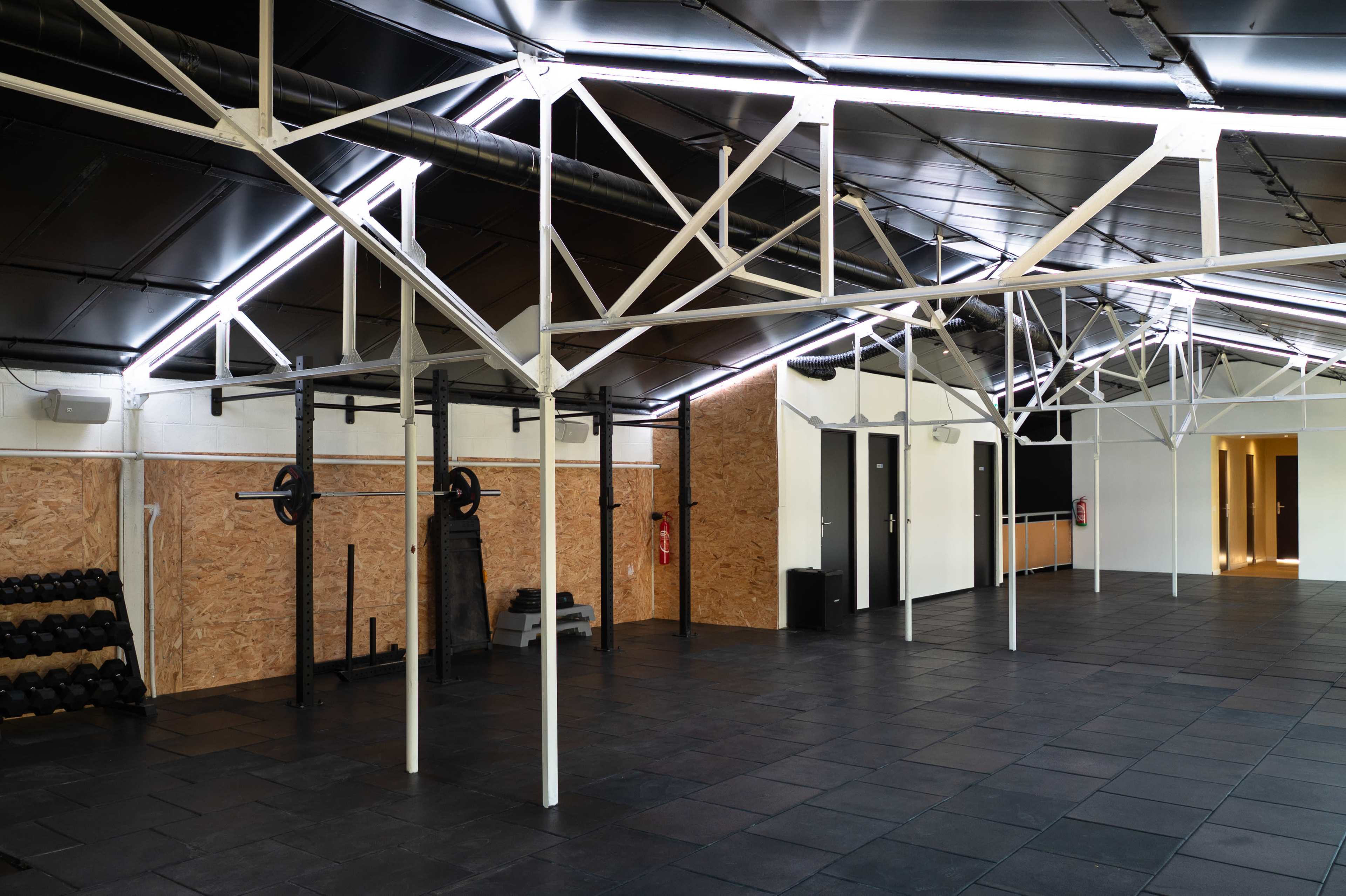 The image shows a spacious gym with a black floor, white metal beams, and an arrangement of weights and fitness equipment against the walls.