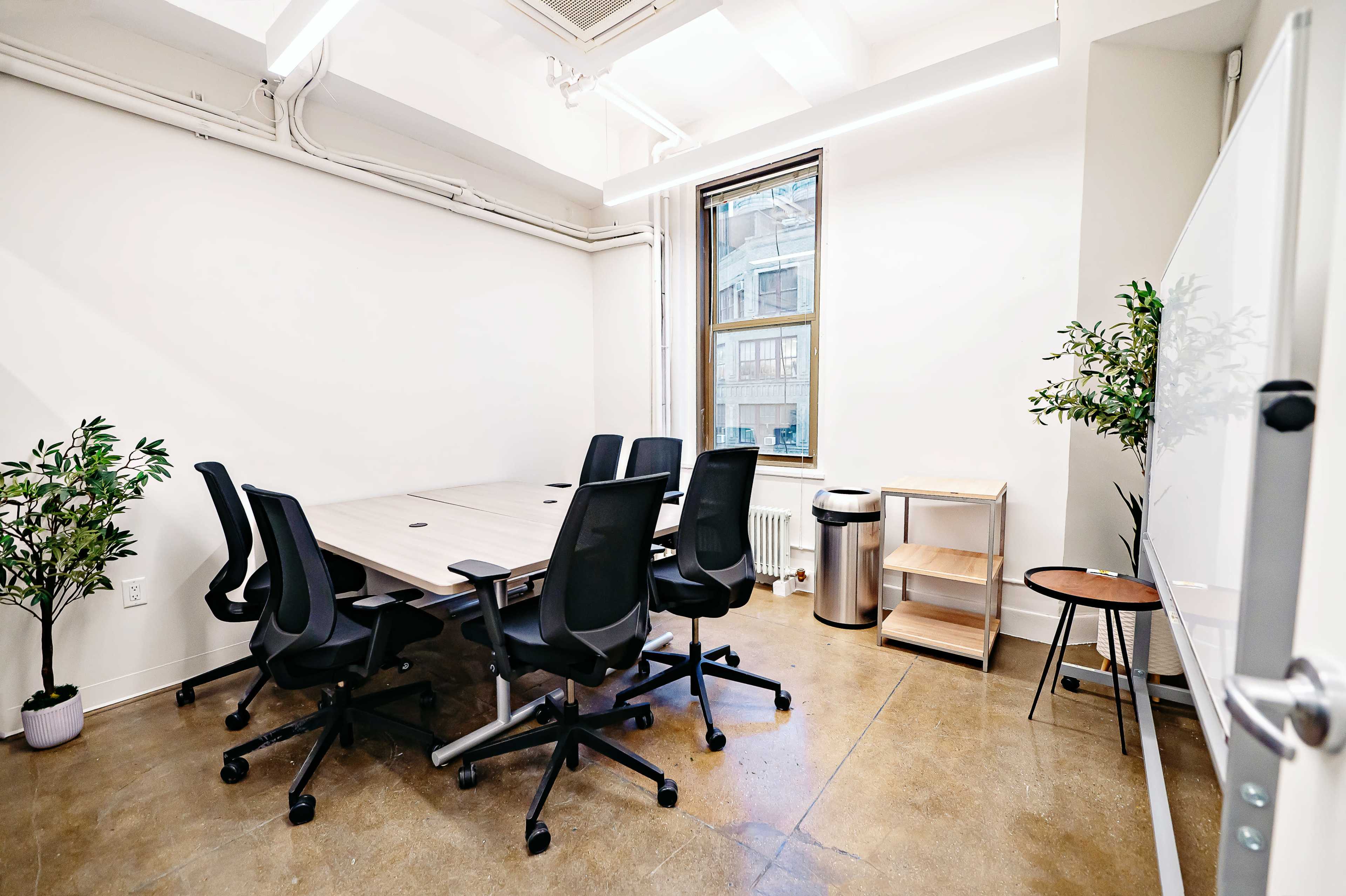 A modern conference room features a large table surrounded by six black office chairs, with a window, a small plant, and a waste bin in the corner.