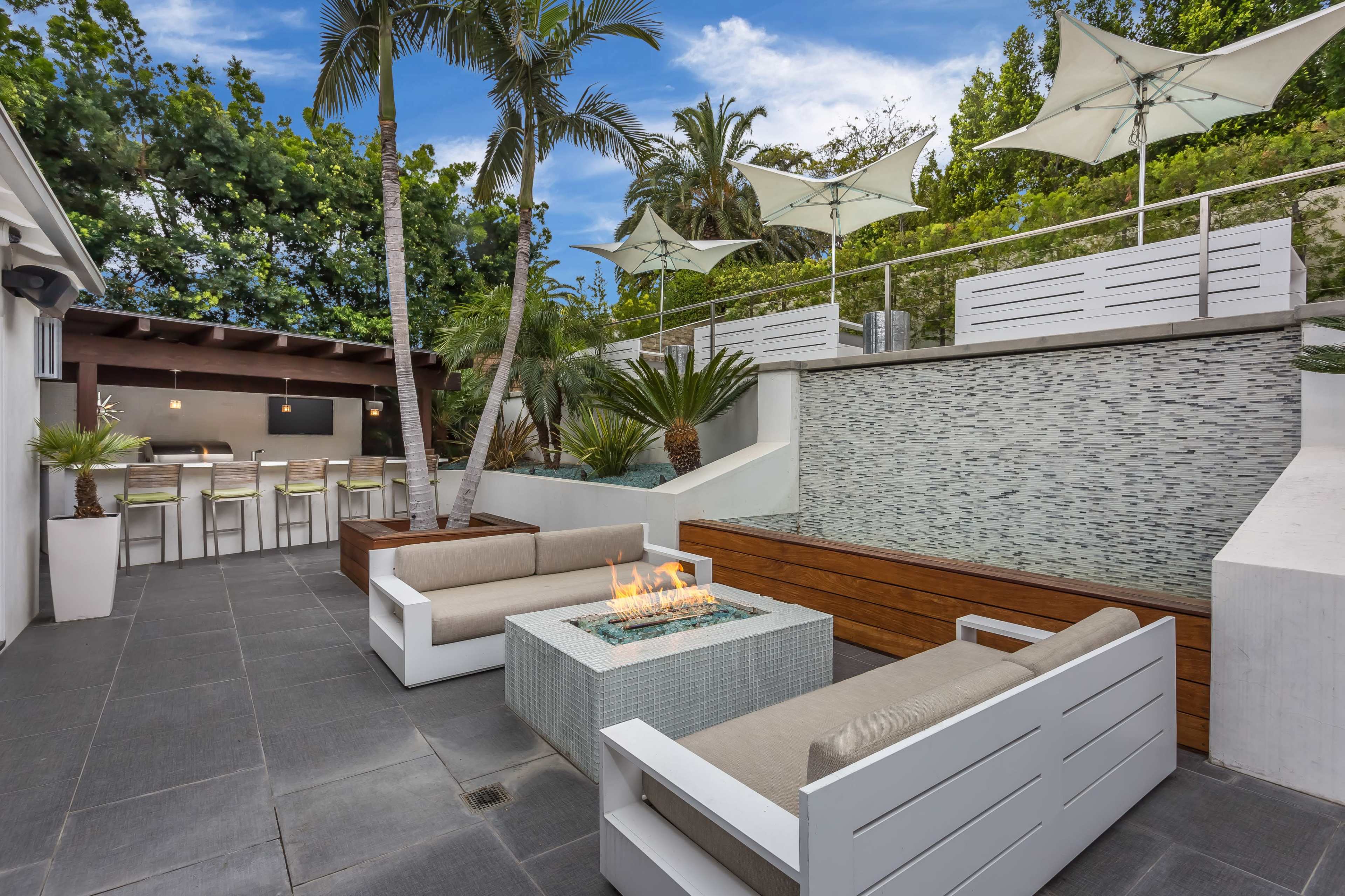The image shows a modern outdoor living space featuring a fire pit, lounge seating, and a water feature, surrounded by greenery and shaded by patio umbrellas.