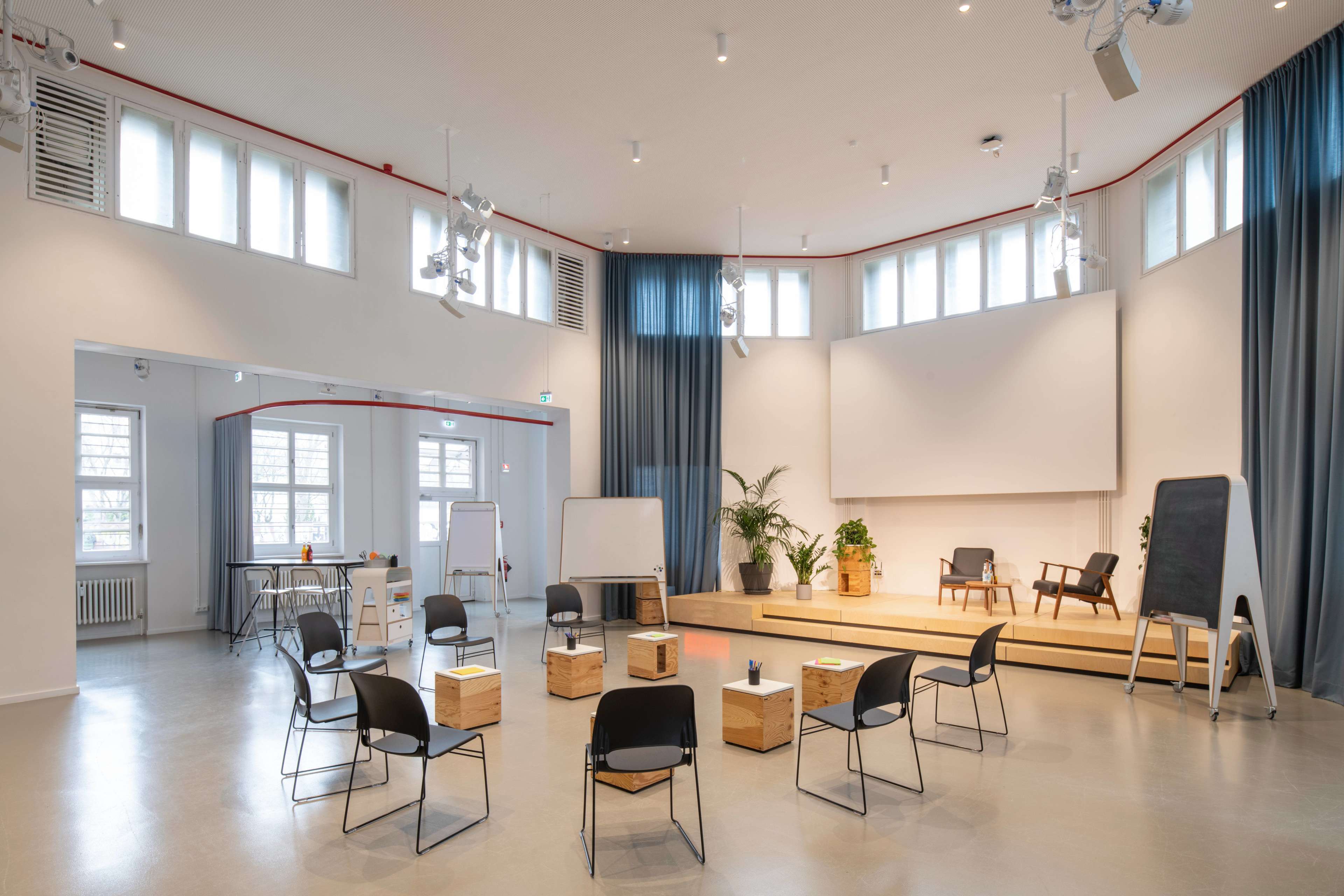 A modern seminar room with several chairs arranged in a circle around wooden boxes, and a large screen on a raised platform at the front.