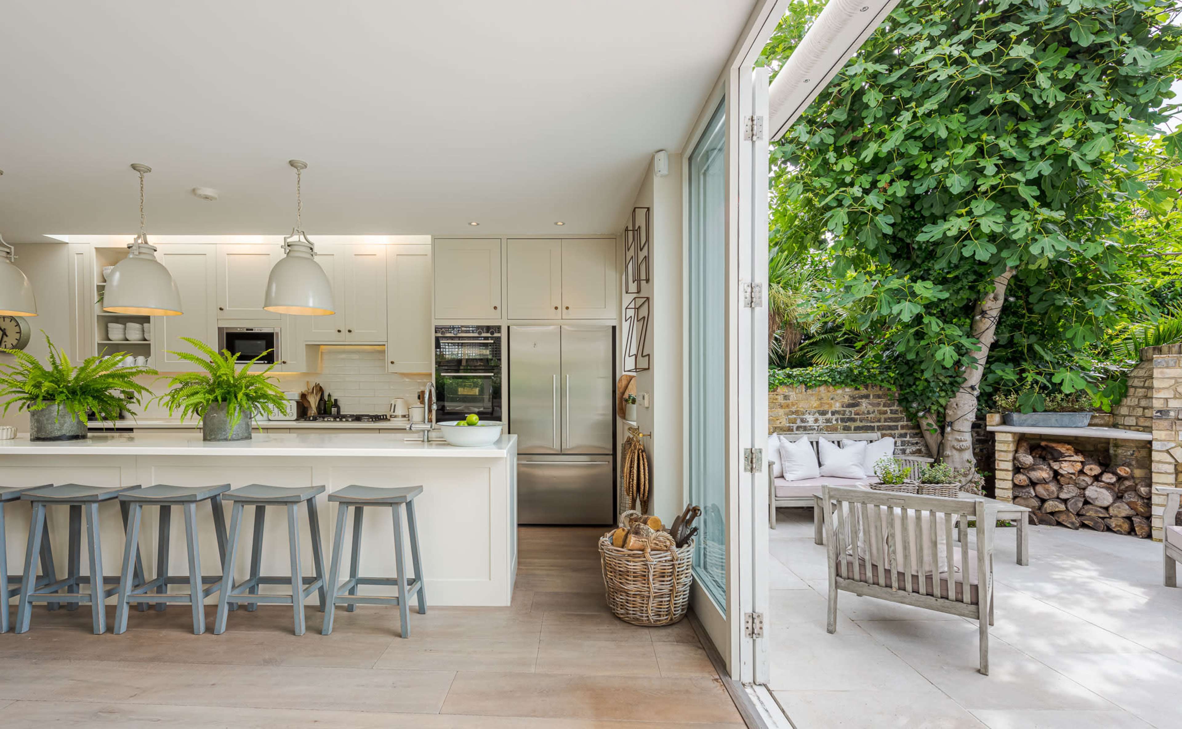 A modern kitchen with a large island, bar stools, and an open view into a garden area featuring a seating arrangement and stacked firewood.