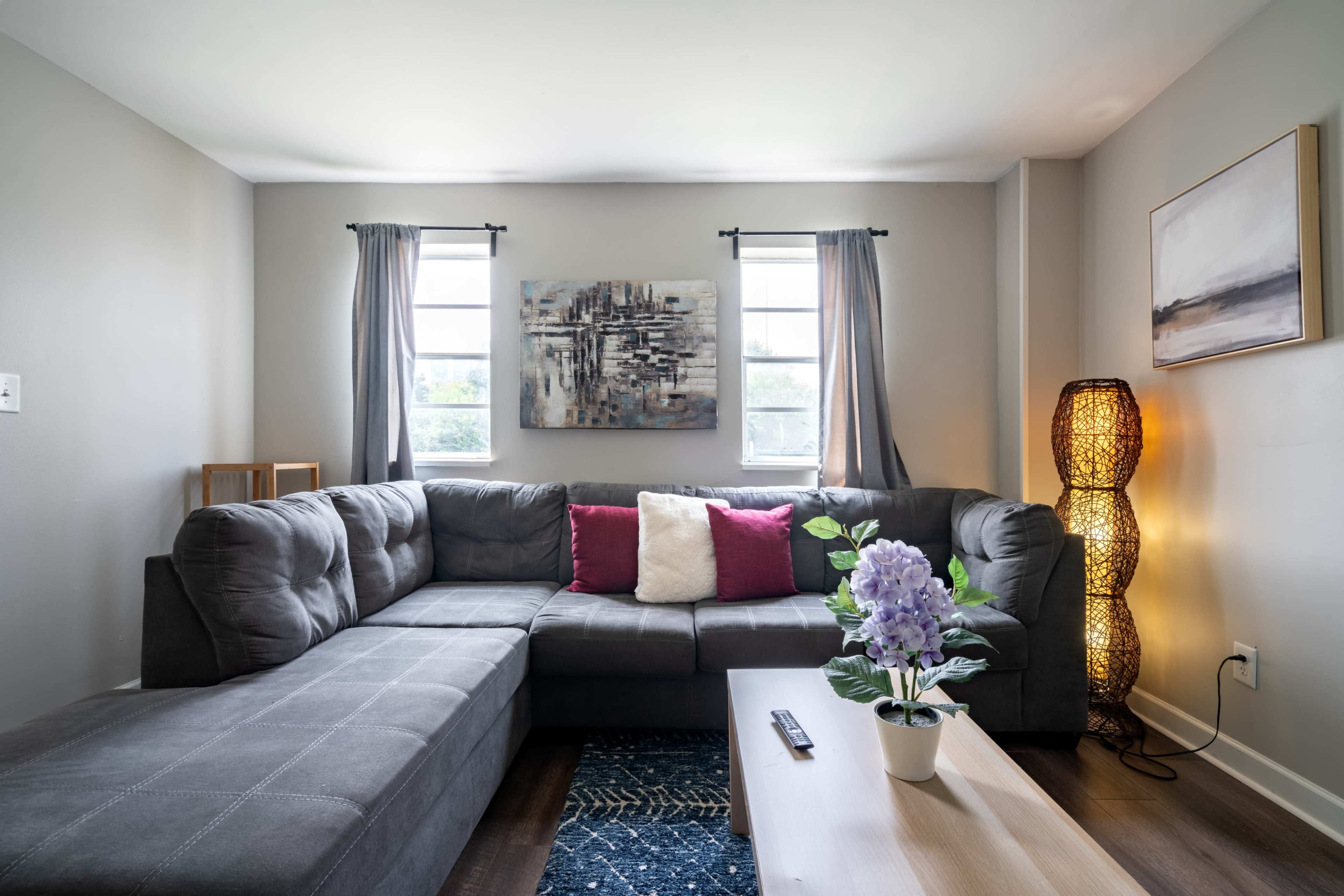 A gray sectional couch with colorful throw pillows is arranged in a bright living room featuring two windows, artwork on the wall, and a coffee table with a plant.