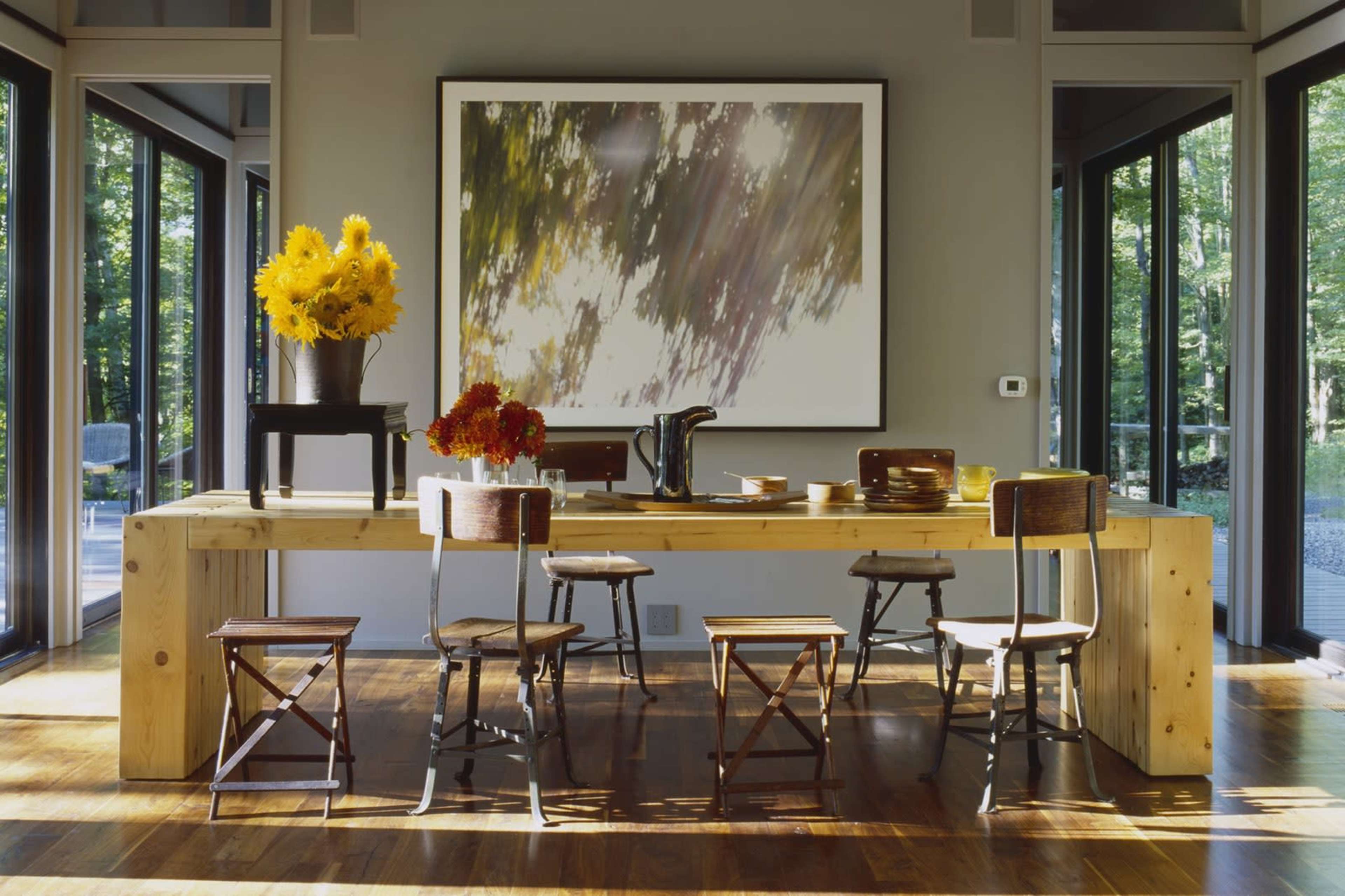 A modern dining area features a large wooden table surrounded by several wooden chairs, with a vibrant flower arrangement on the table and large windows allowing natural light.