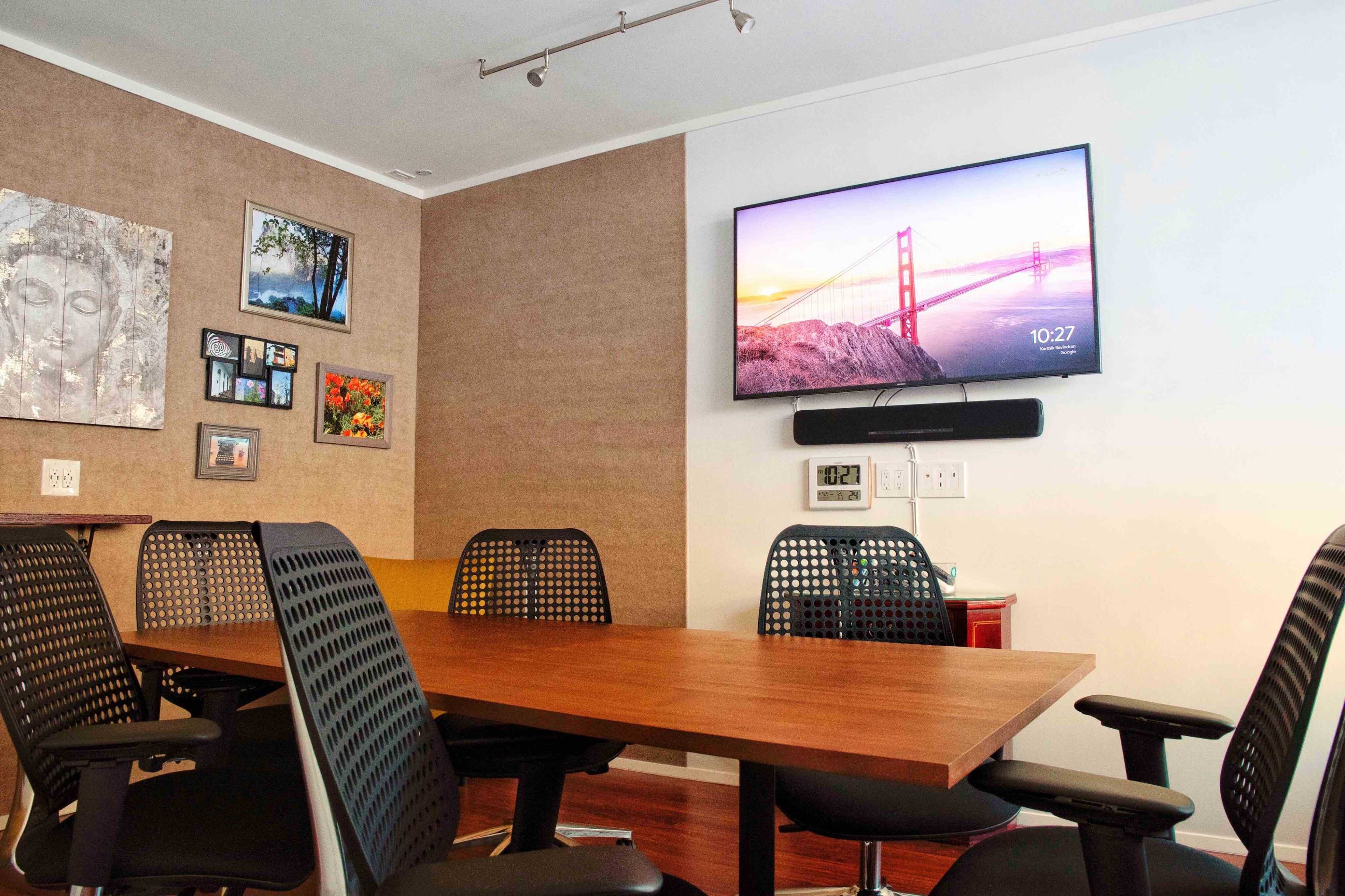 A modern conference room features a wooden table surrounded by black mesh chairs, with a television displaying an image of the Golden Gate Bridge on the wall.