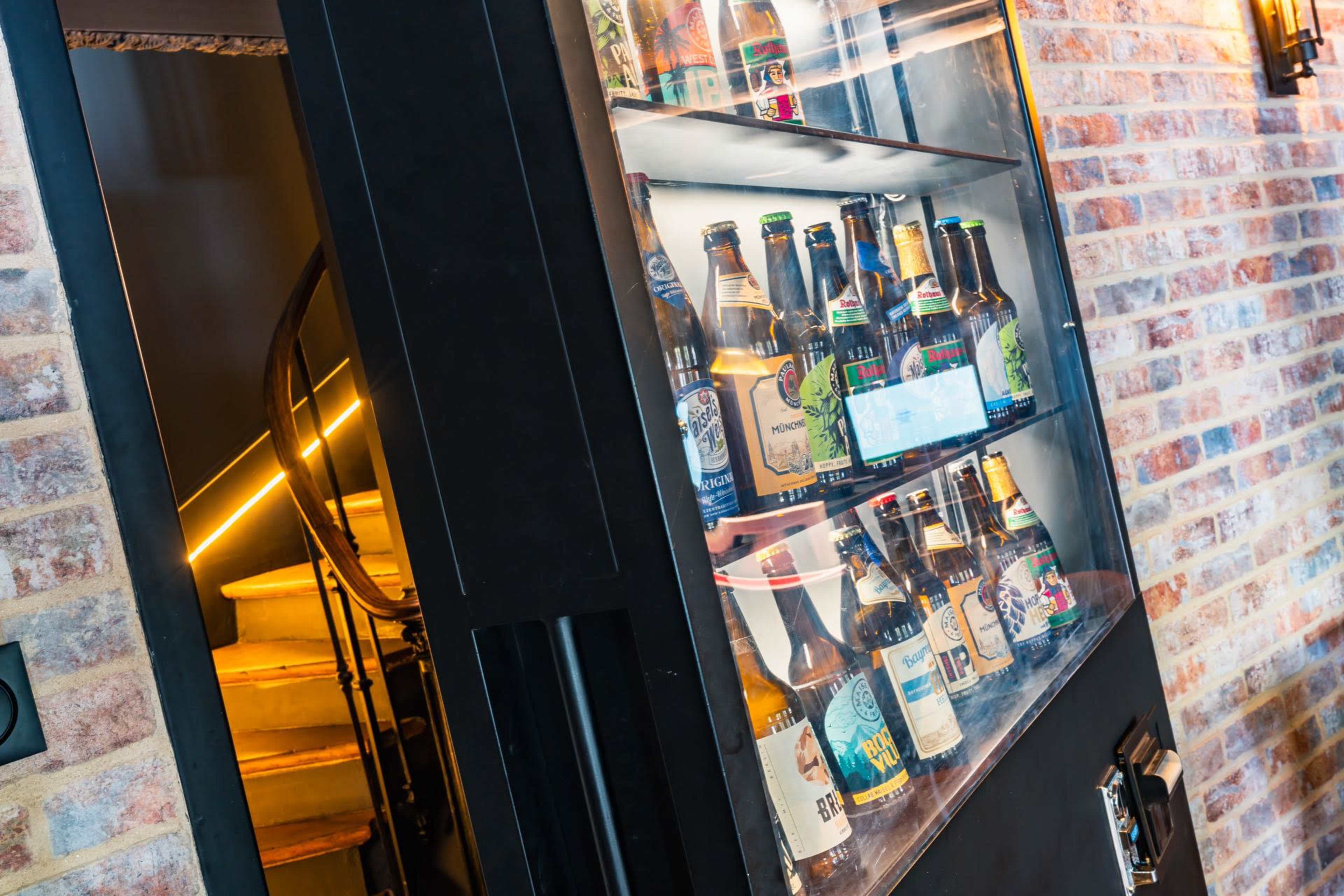 A vending machine filled with various bottled beers is positioned next to a staircase with wooden steps.