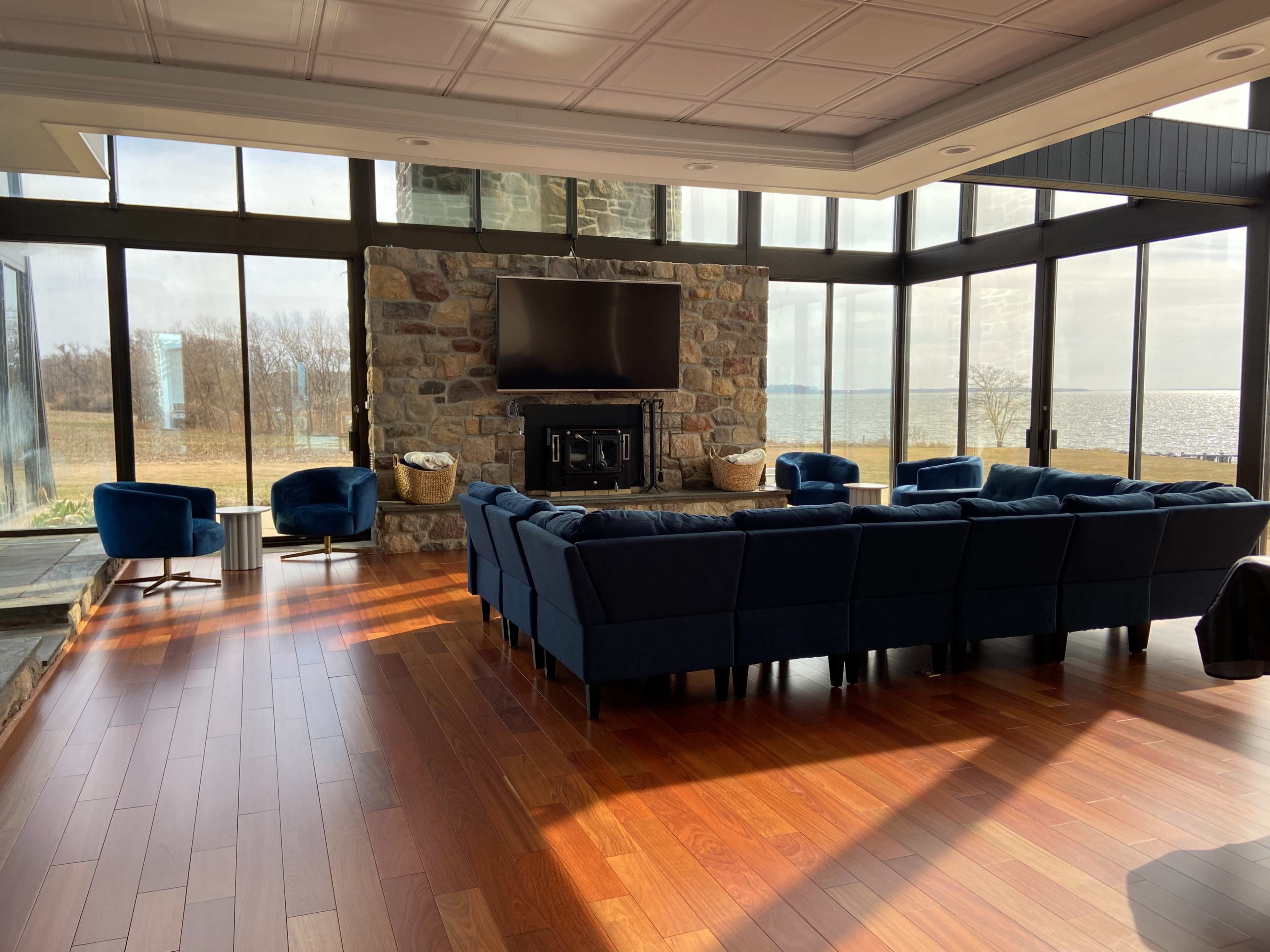 The room features a large, sectional navy blue sofa arranged around a stone fireplace and a flat-screen television, with large windows providing a view of the outdoors.