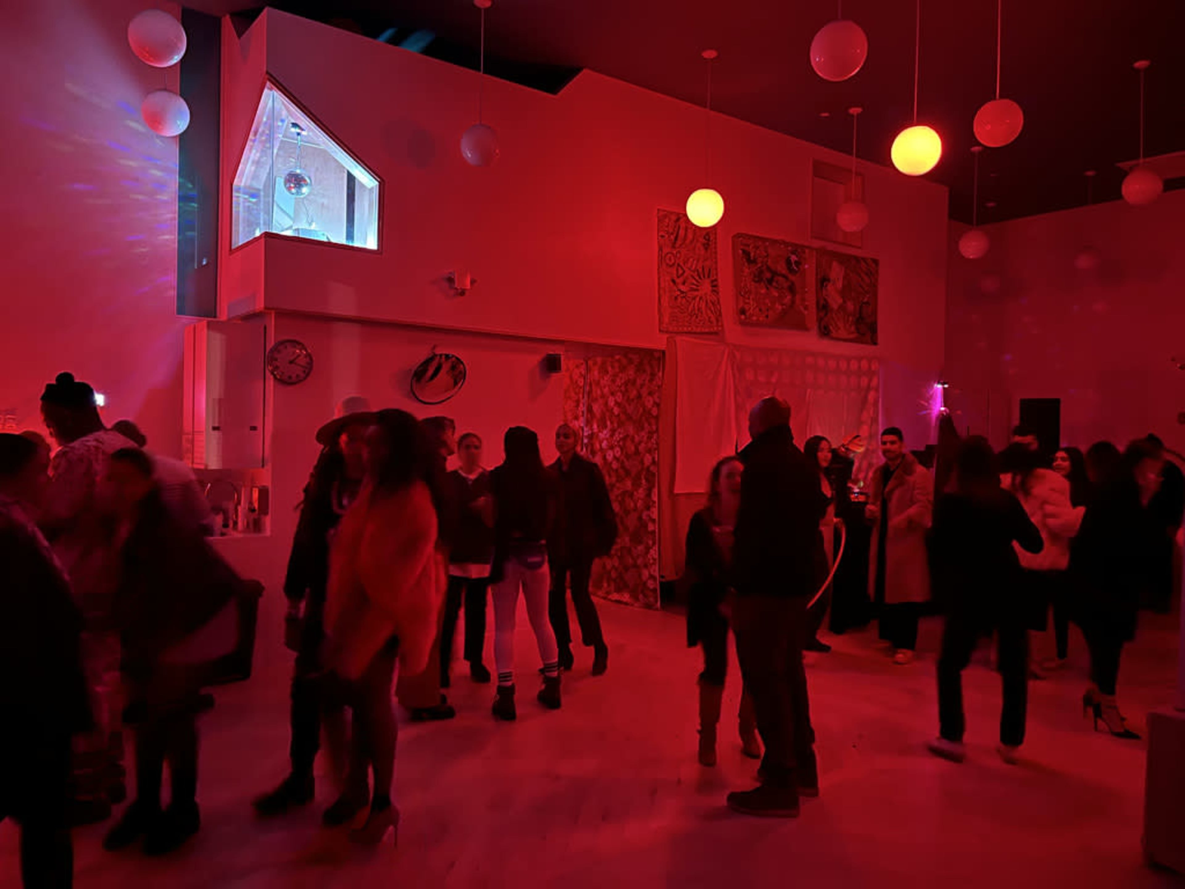A crowd of people socializes and dances in a brightly lit room with red lighting and colorful decorations.