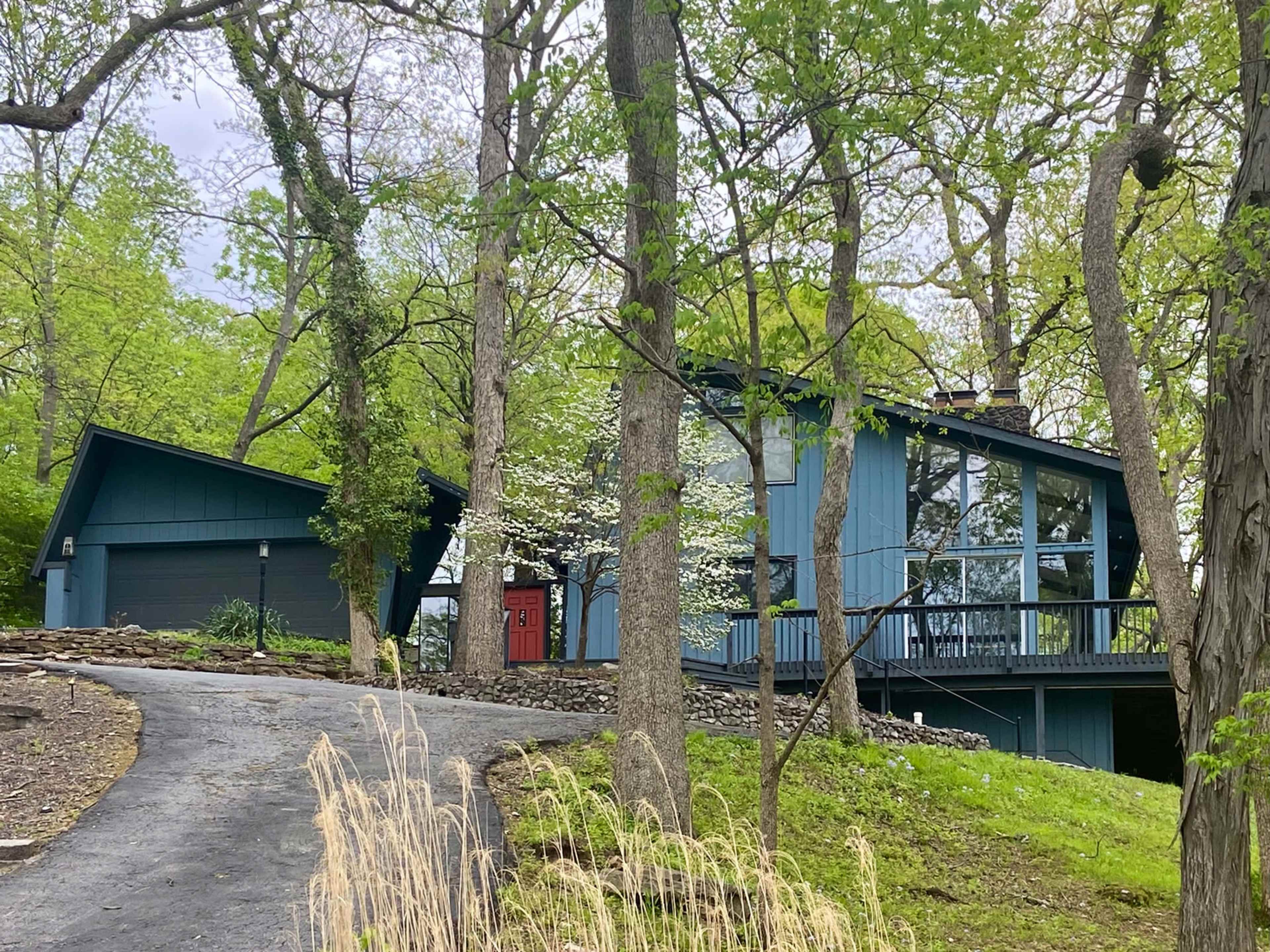 A modern blue house with a gabled roof is nestled among trees, featuring large windows and a red front door, situated at the end of a gravel driveway.