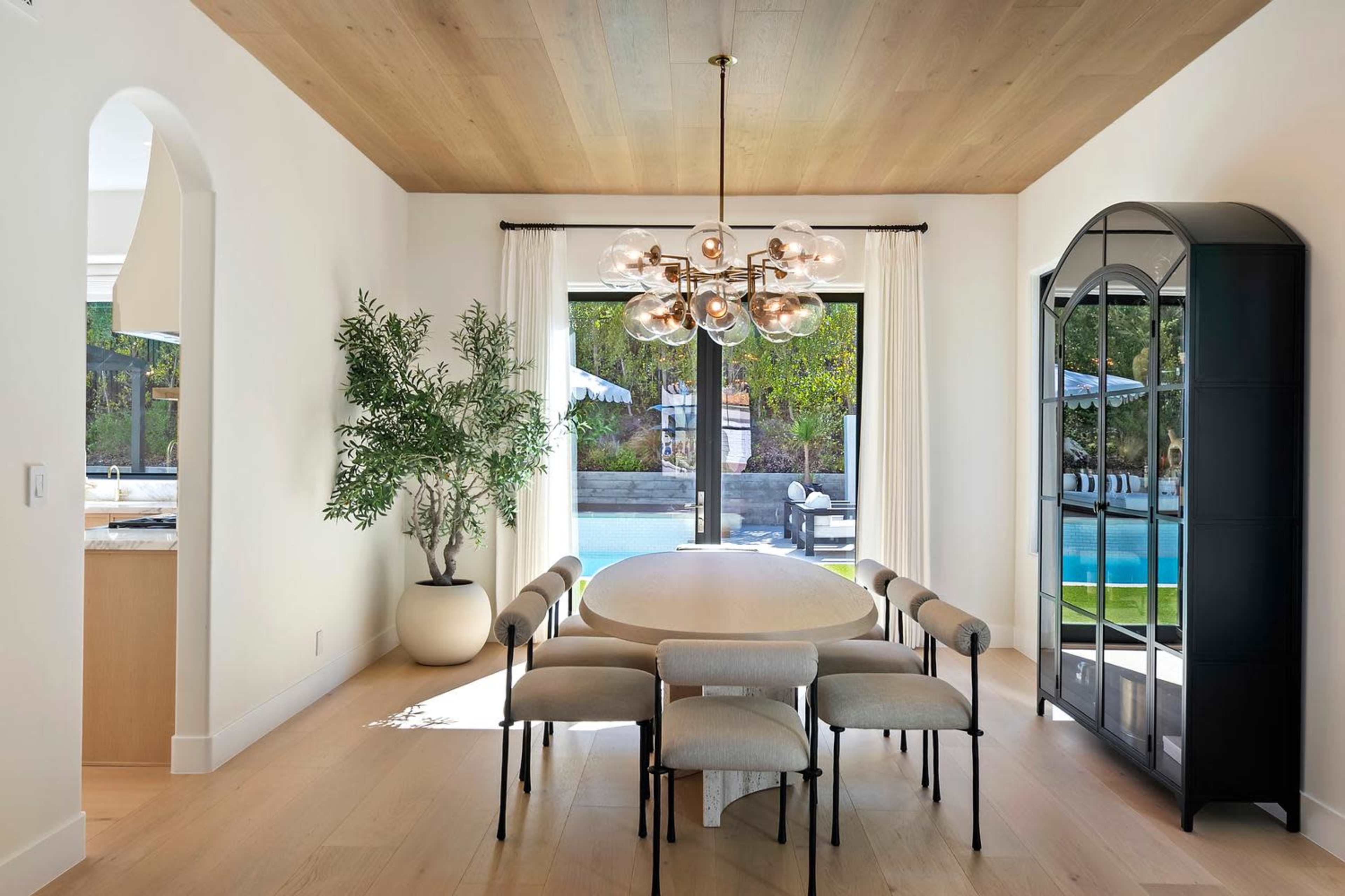 A modern dining area features a round table with upholstered chairs, a large chandelier, and sliding glass doors that open to a poolside view.