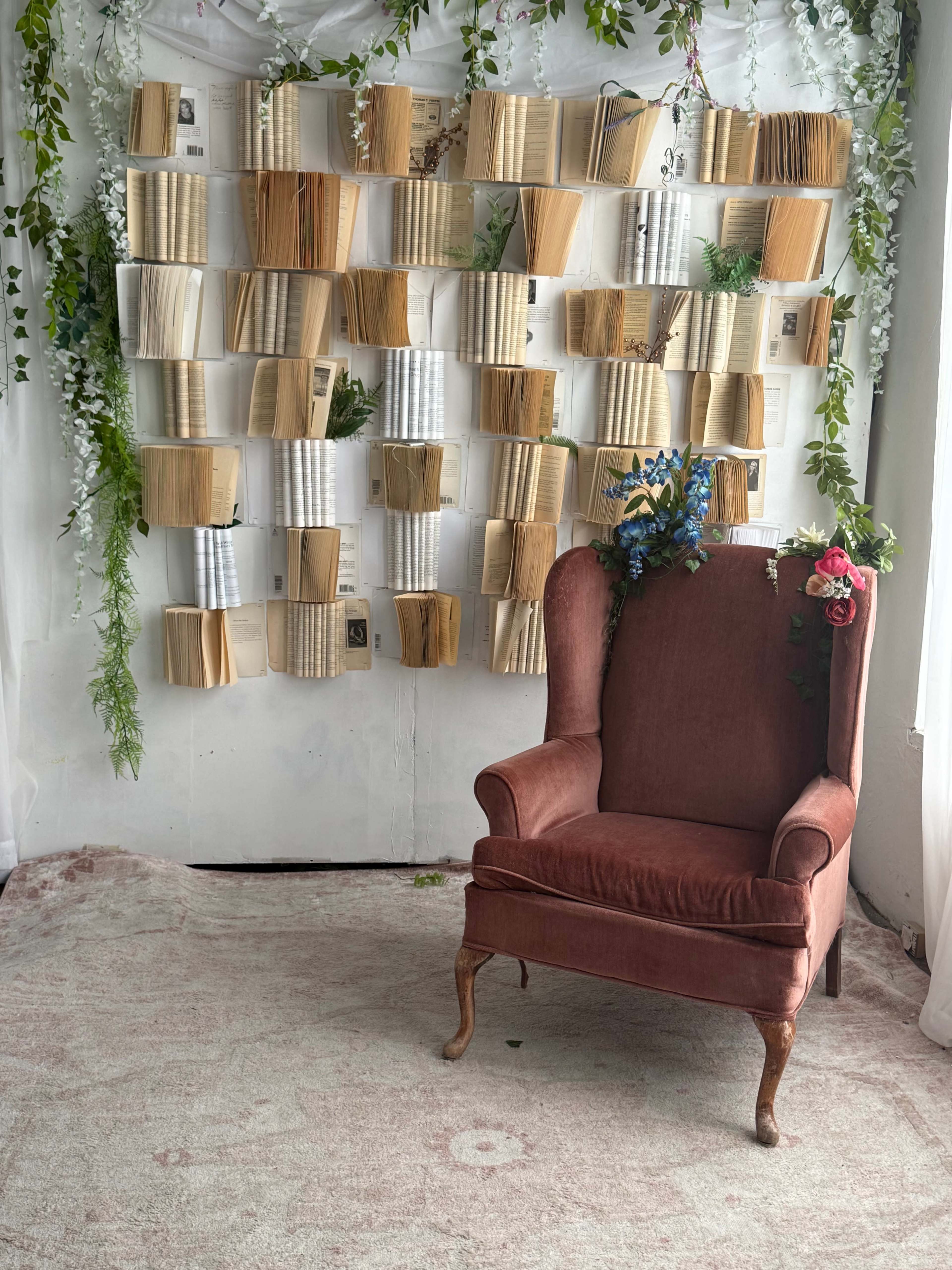 A vintage red armchair sits in front of a decorative wall made of open books and greenery.