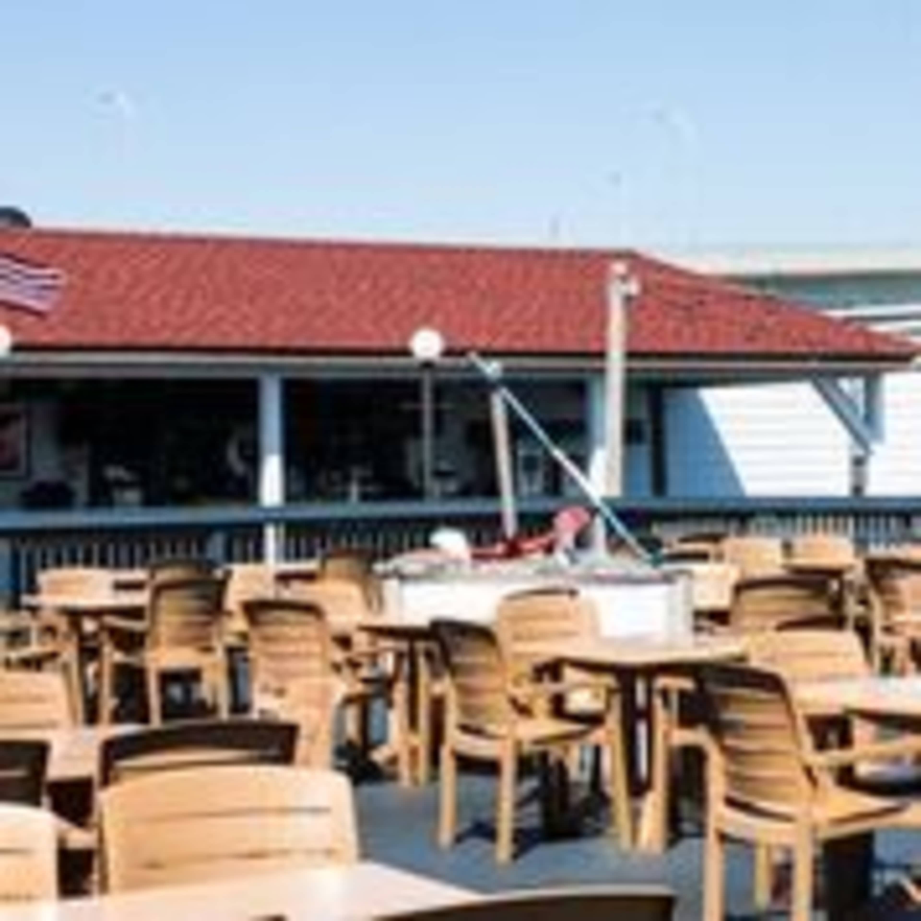 An outdoor dining area with wooden tables and chairs near a building with a red roof, and an American flag flying nearby.
