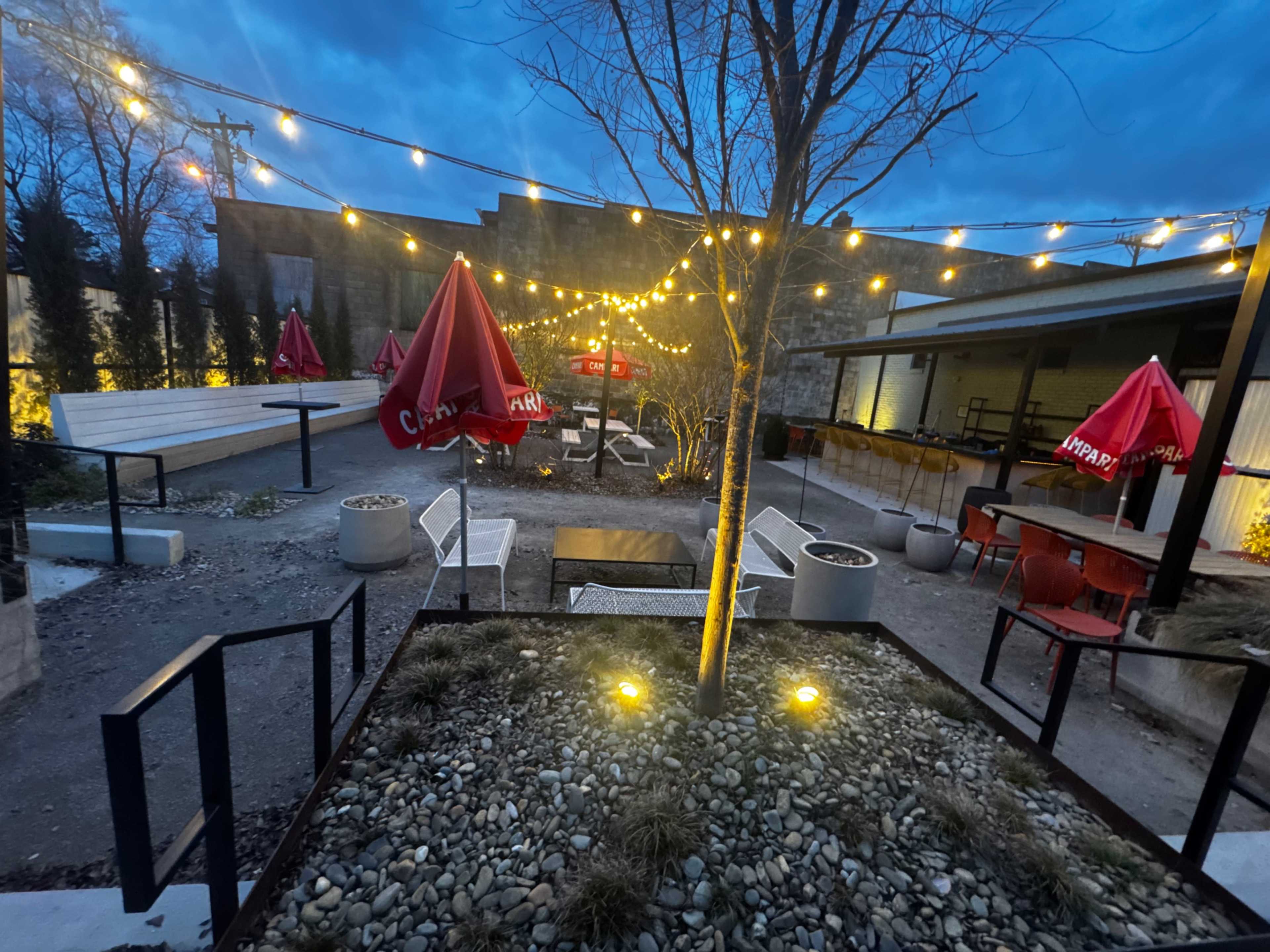 The image shows an outdoor seating area with string lights, red umbrellas, and modern furniture amid gravel and planters.