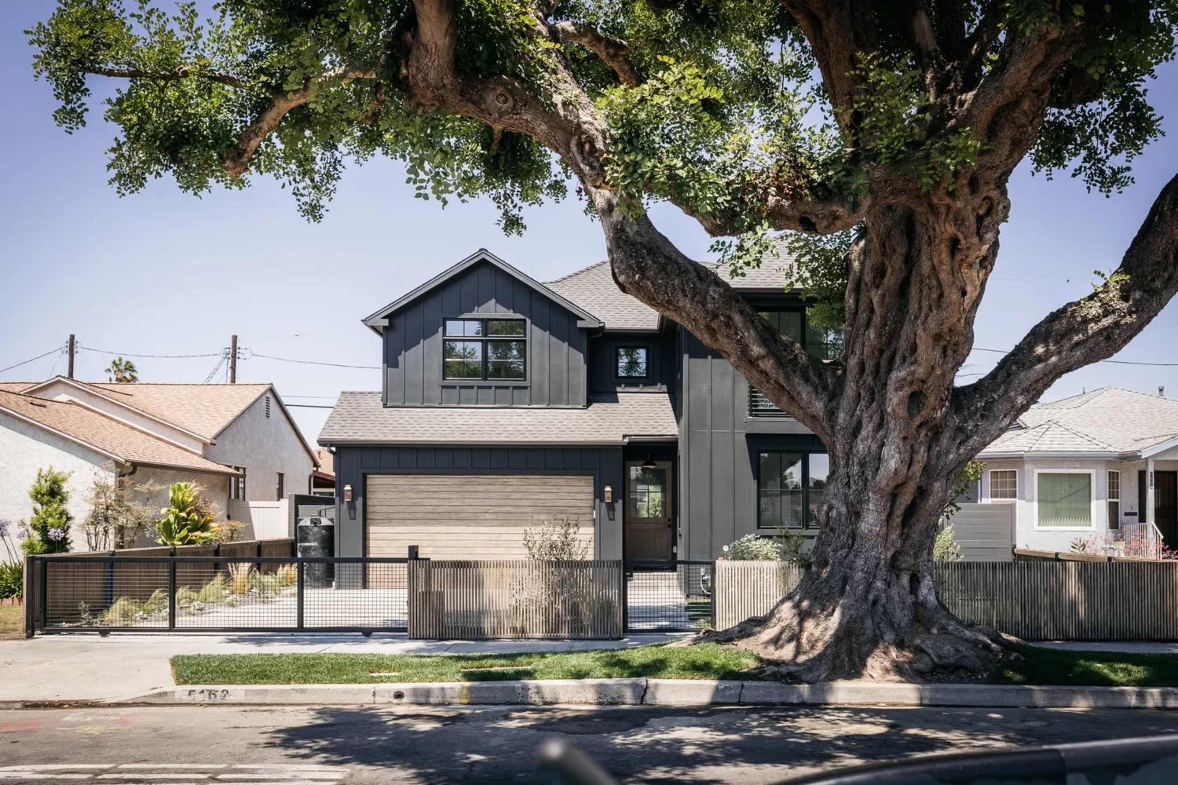 A modern two-story house with a dark exterior and large windows stands next to a massive tree, surrounded by a fence and well-kept landscaping.