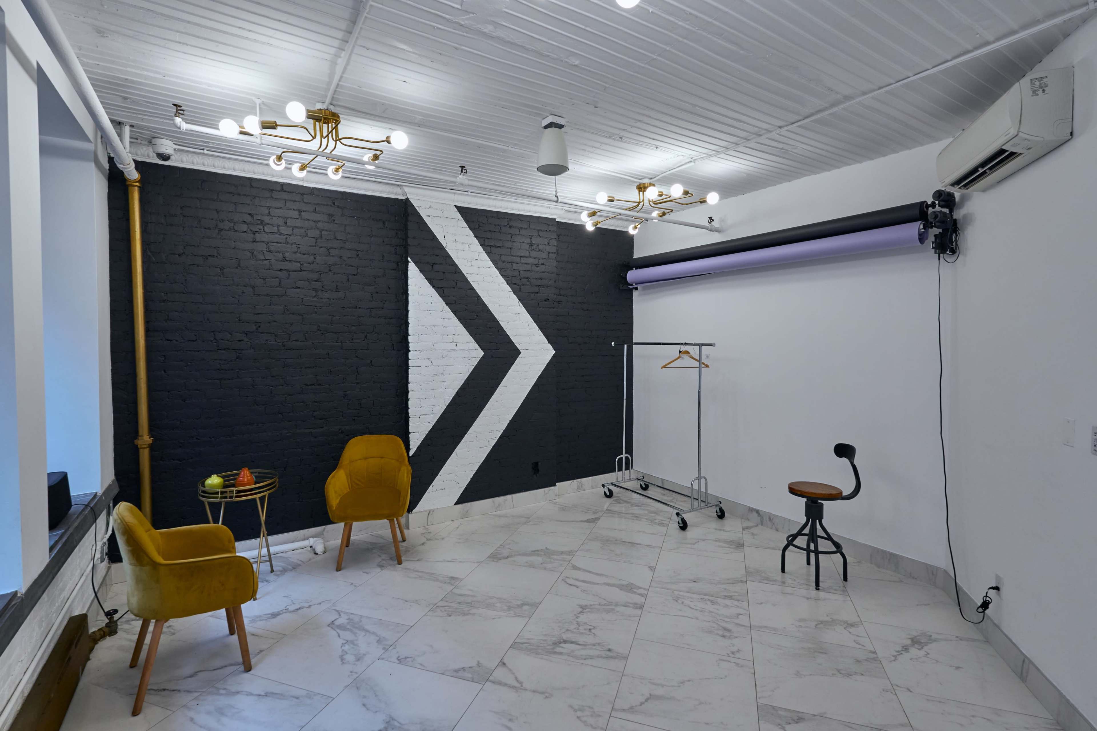 A spacious studio with marble flooring, featuring two yellow chairs, a clothing rack, a stool, and a black and white accent wall with a geometric design.