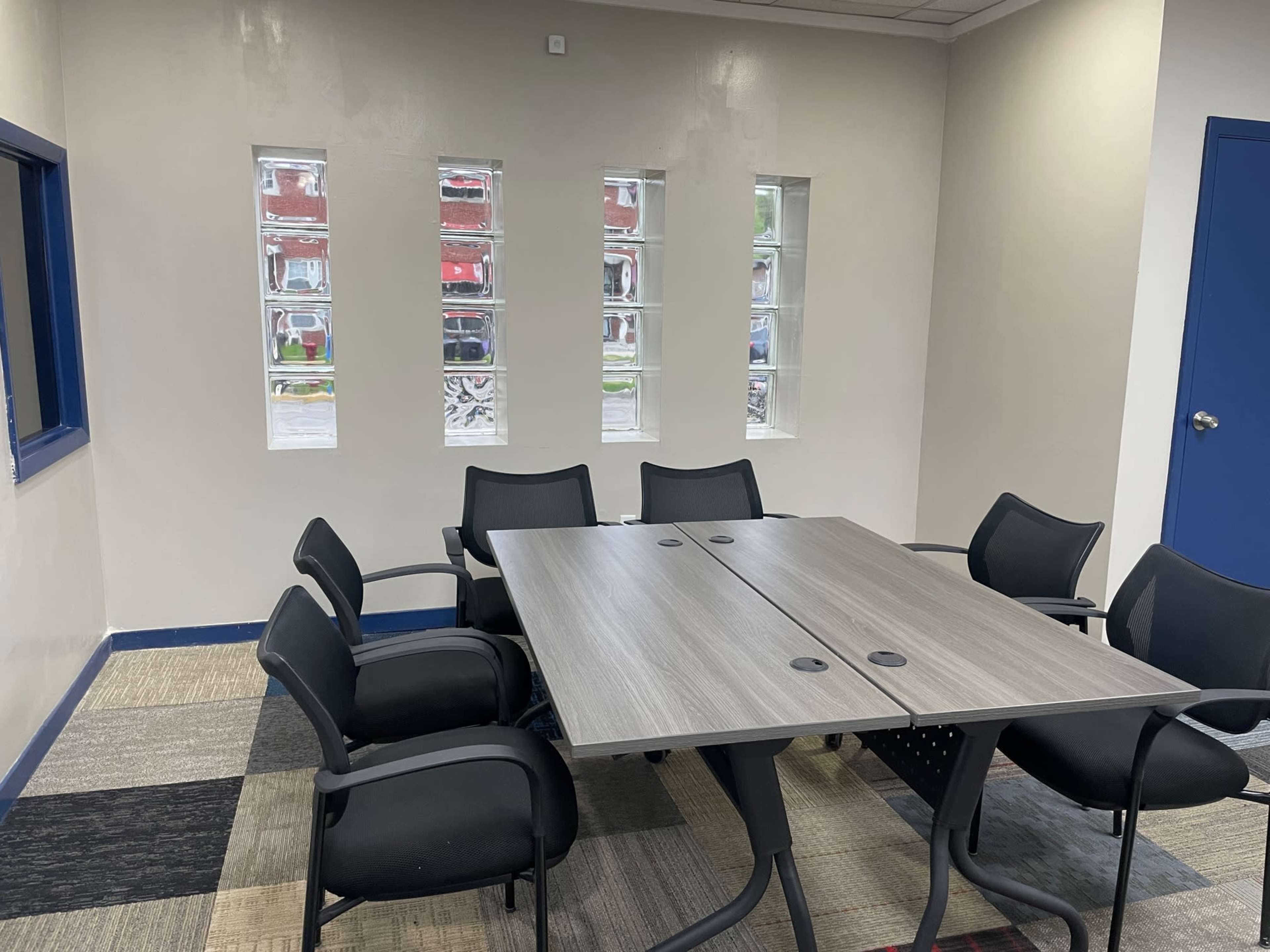 A conference room features a rectangular table surrounded by six black chairs and has three narrow windows along one wall.