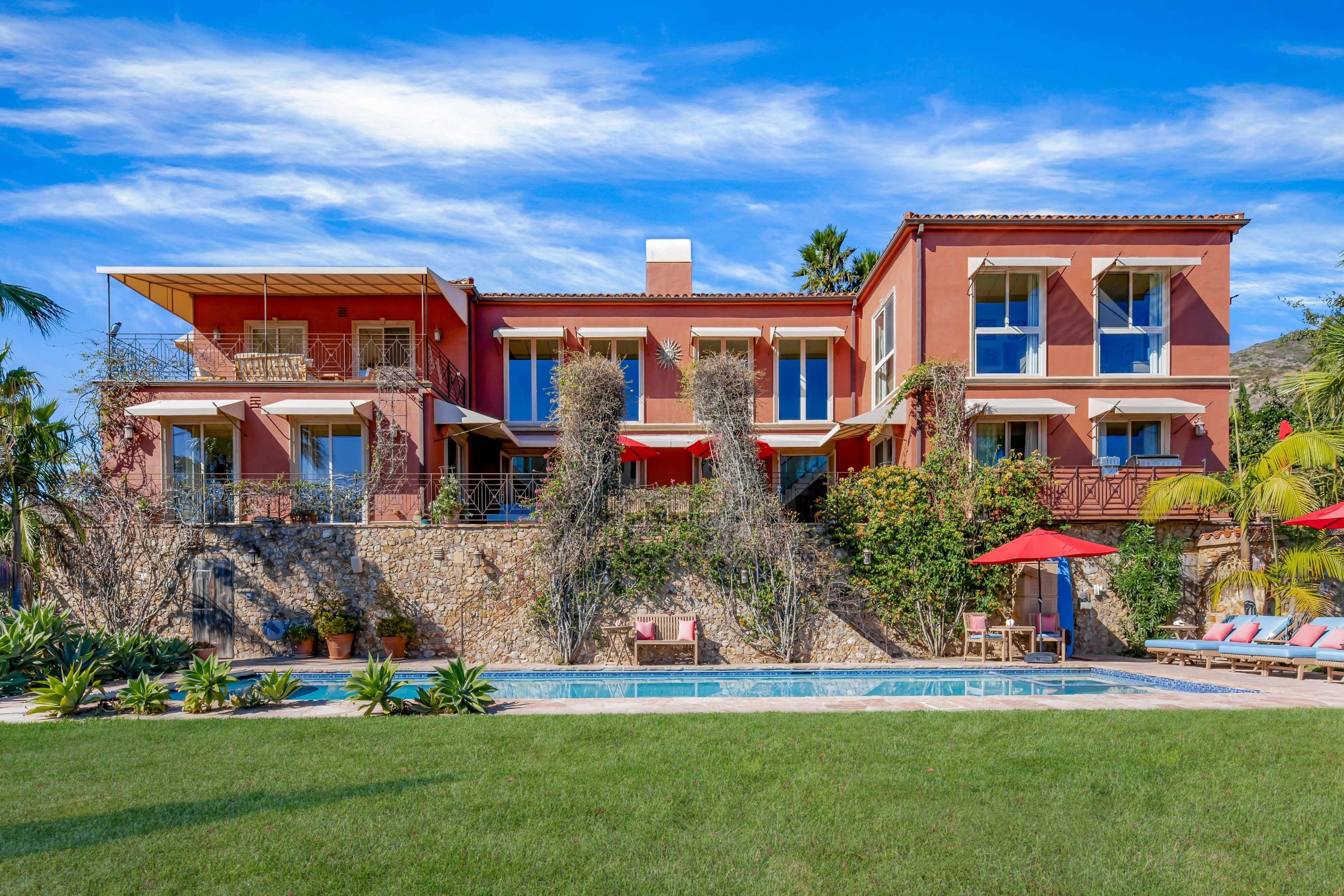 A large, multi-level red villa with balconies sits alongside a swimming pool and a green lawn, surrounded by palm trees and vibrant landscaping.