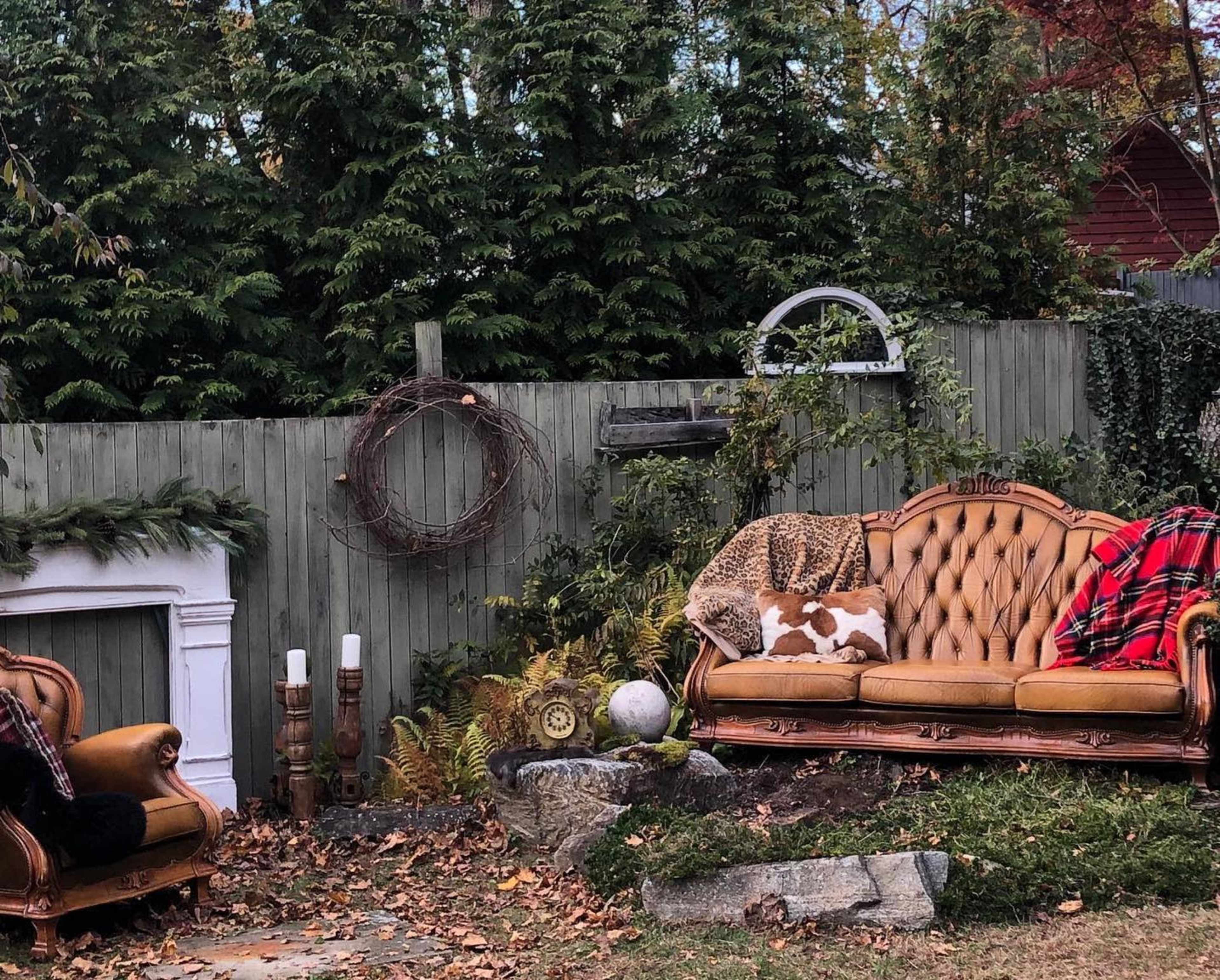 An outdoor seating area with a brown velvet sofa, a matching armchair, and various decorative elements against a wooden fence.