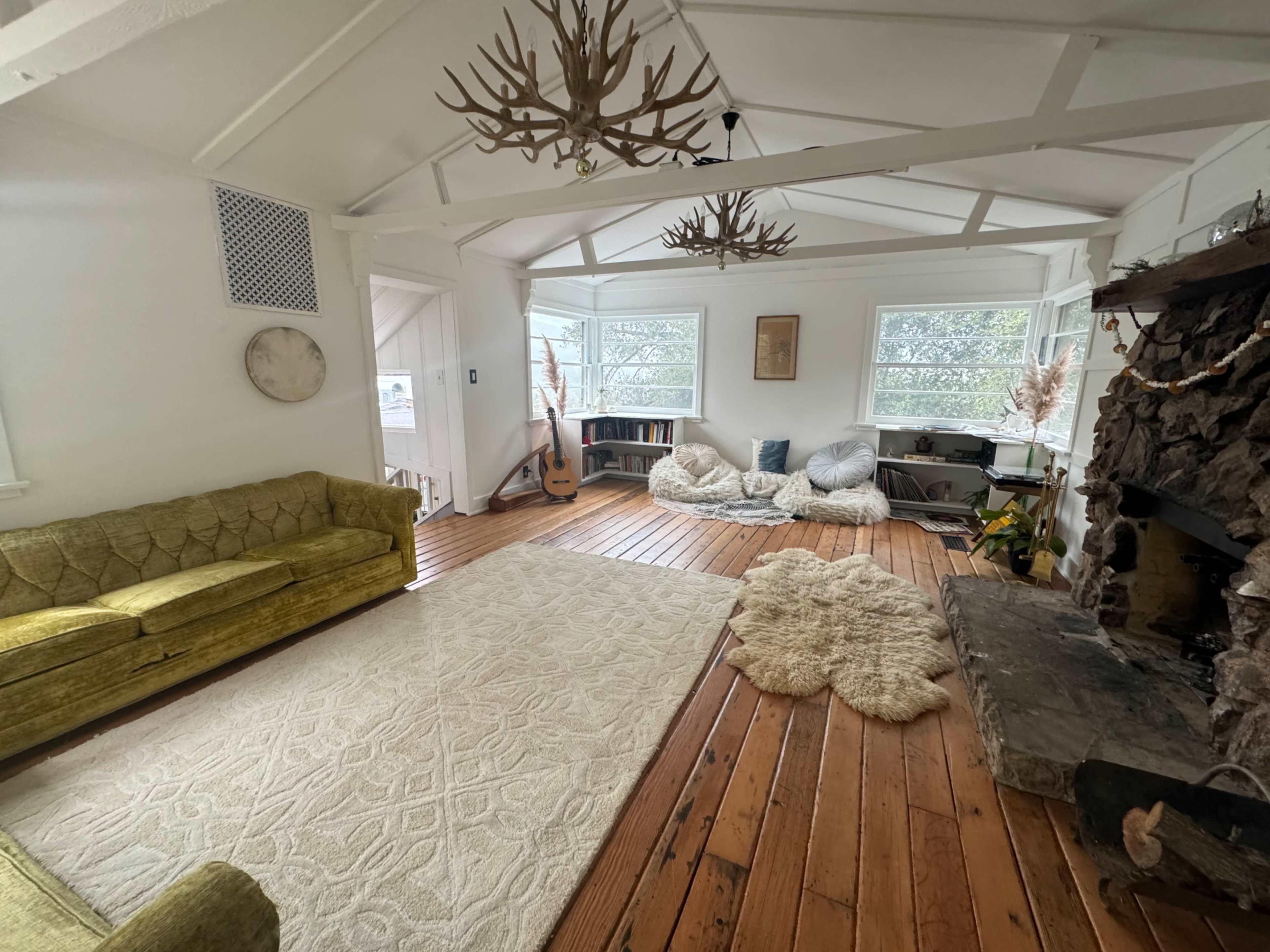 The image shows a cozy living room with a green sofa, large windows, a stone fireplace, and seating areas made of soft cushions and rugs on wooden flooring.