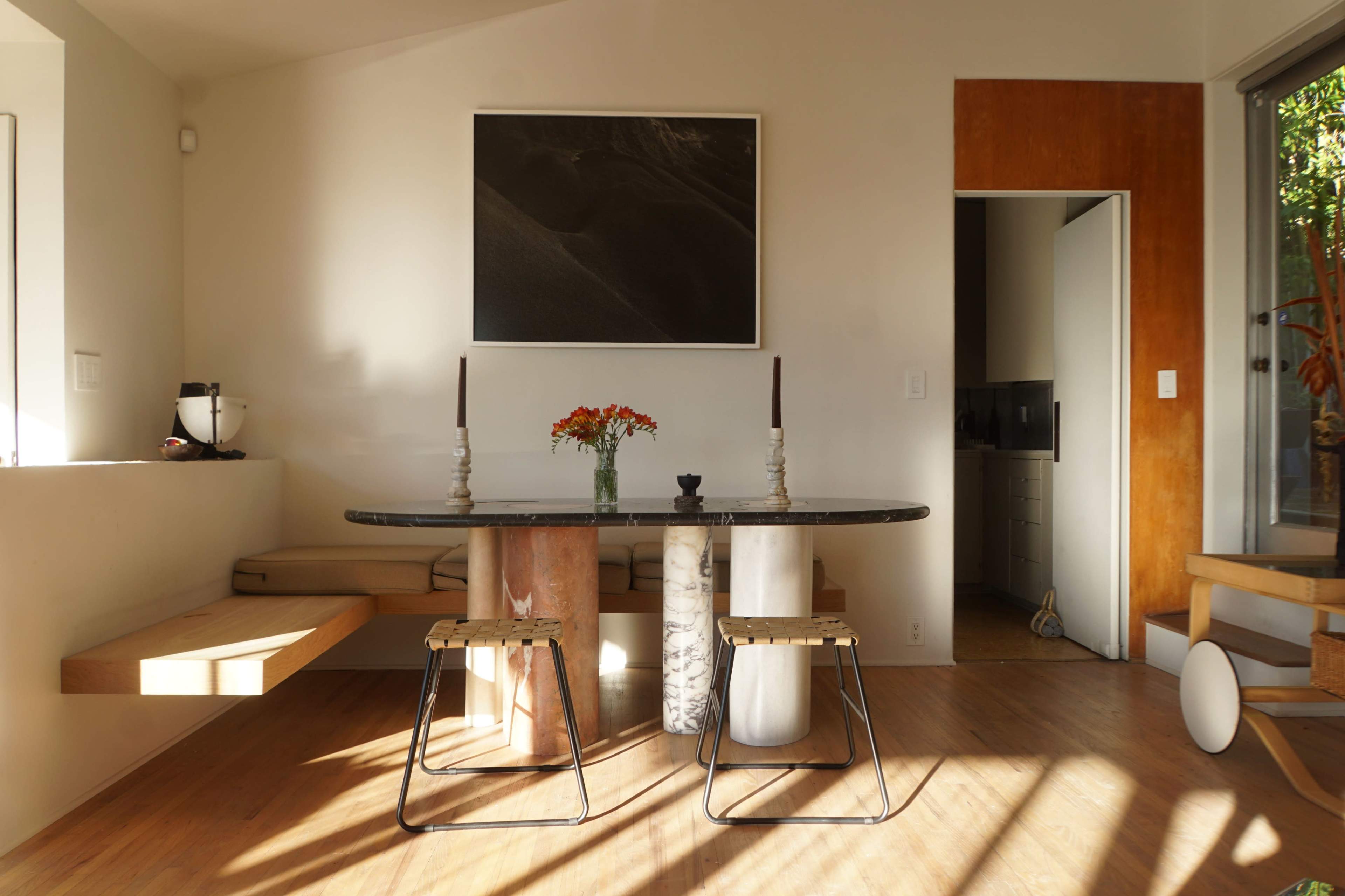A modern dining area features a round table with two stools, a vase of flowers, and a large black-and-white photograph on the wall.