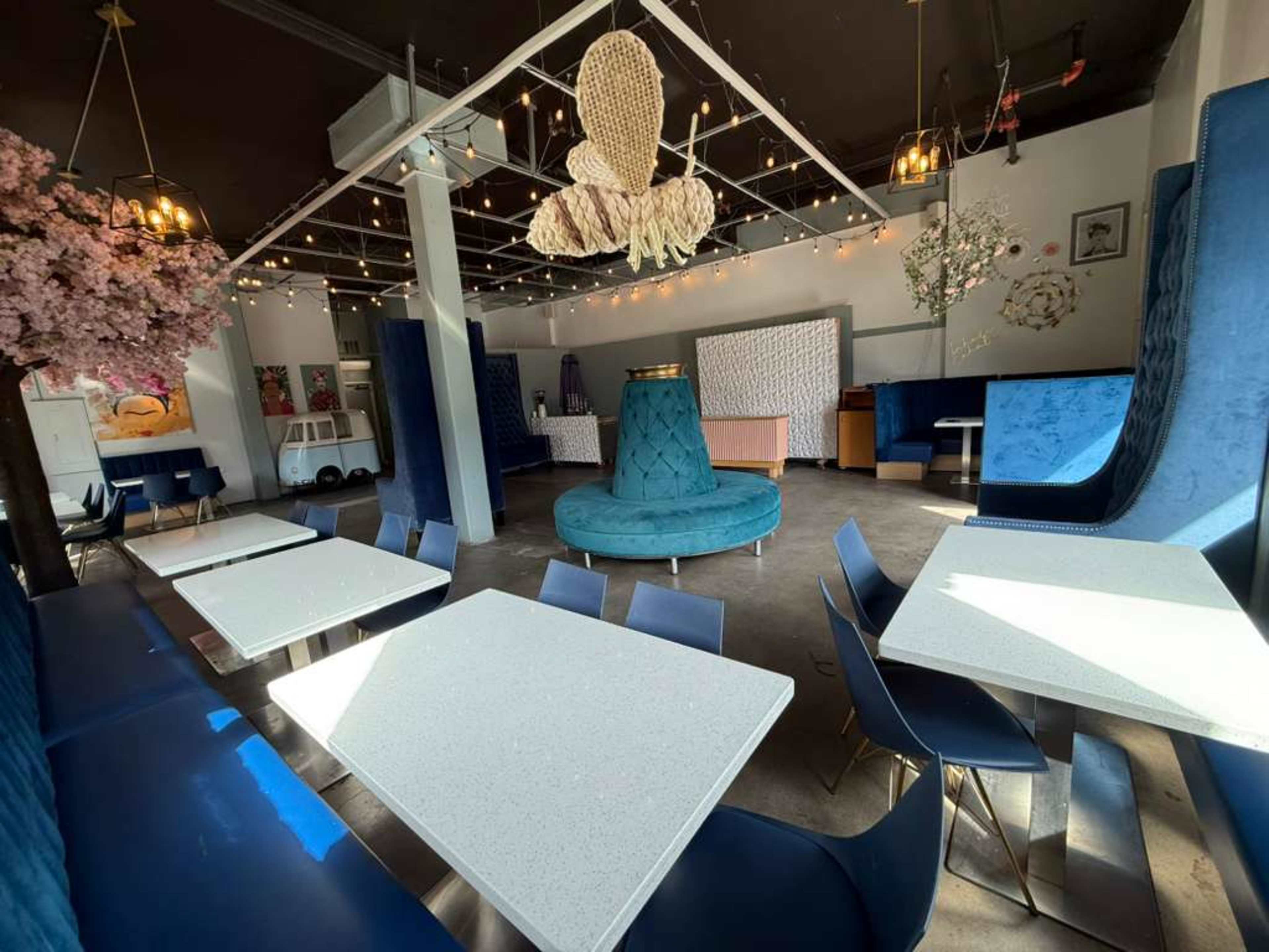 The image shows a brightly lit cafe interior featuring blue velvet seating, white tables, and decorative elements like hanging lights and artificial cherry blossoms.