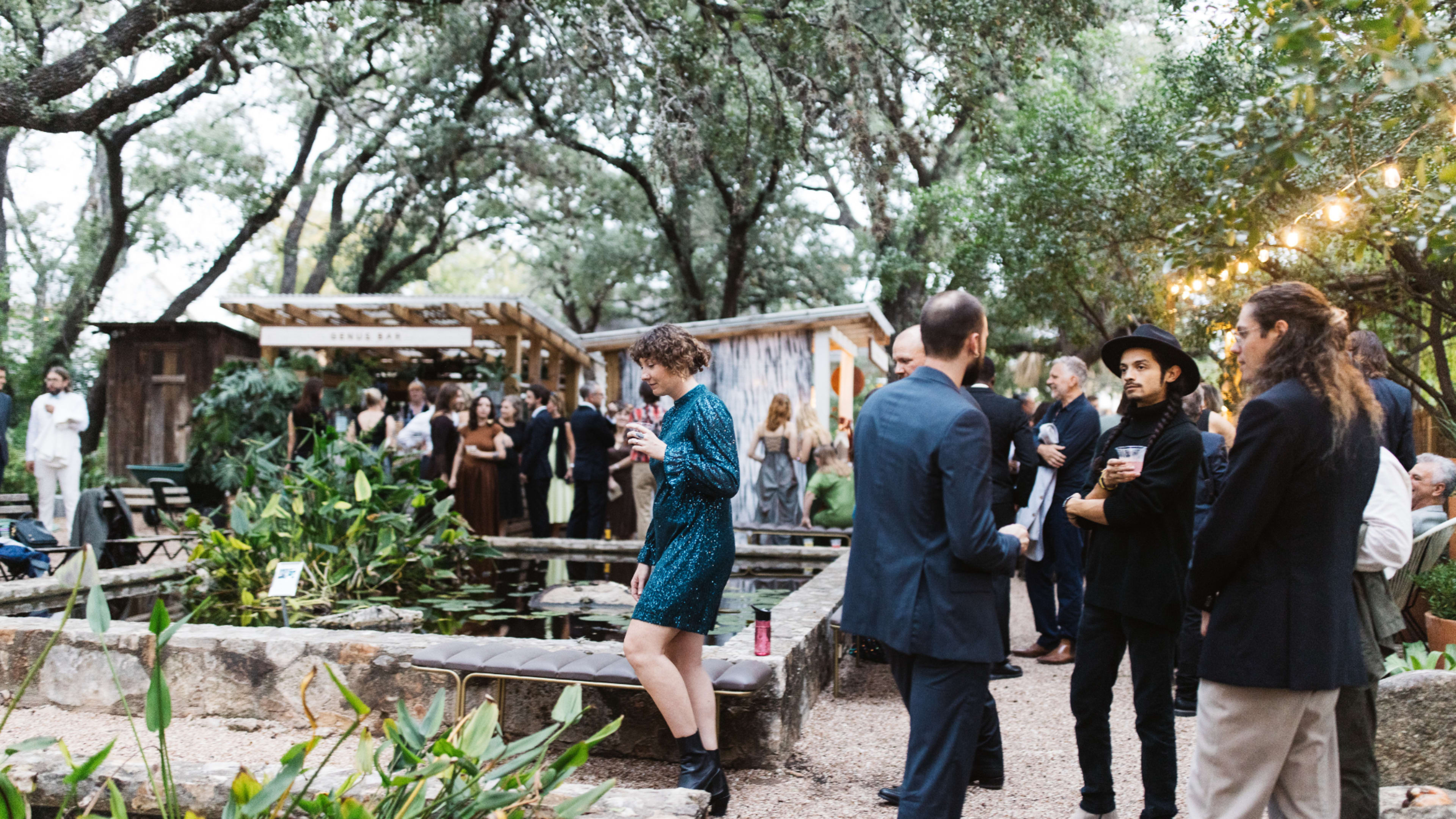 A group of people socialize around a garden pond, surrounded by trees and outdoor decor at a gathering.