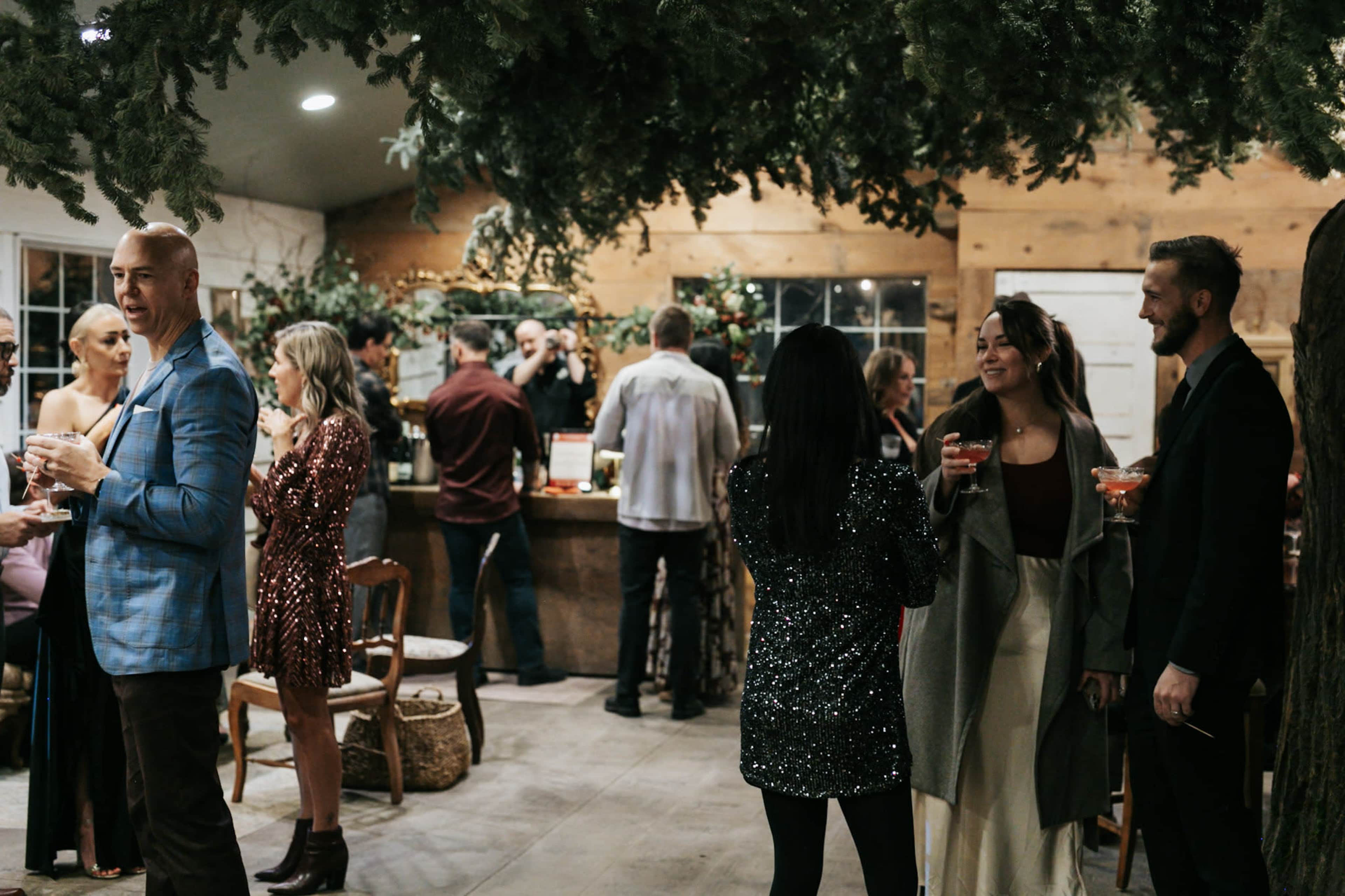 A group of people socialize at a festive indoor gathering decorated with greenery, standing around a bar while enjoying drinks.
