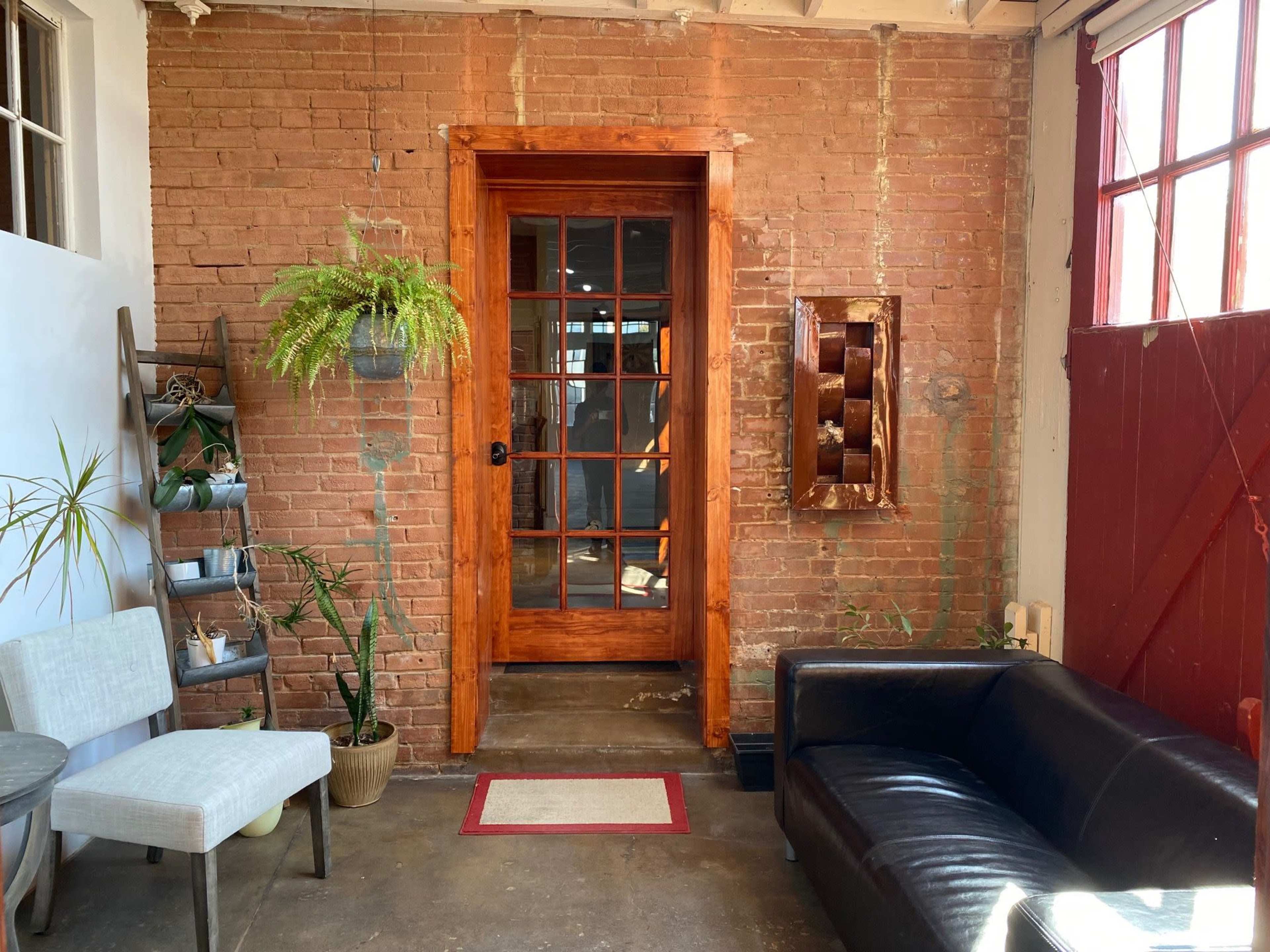 The image shows a brick wall with a wooden door framed by a potted plant and artwork, alongside a black couch and two chairs in a cozy interior space.