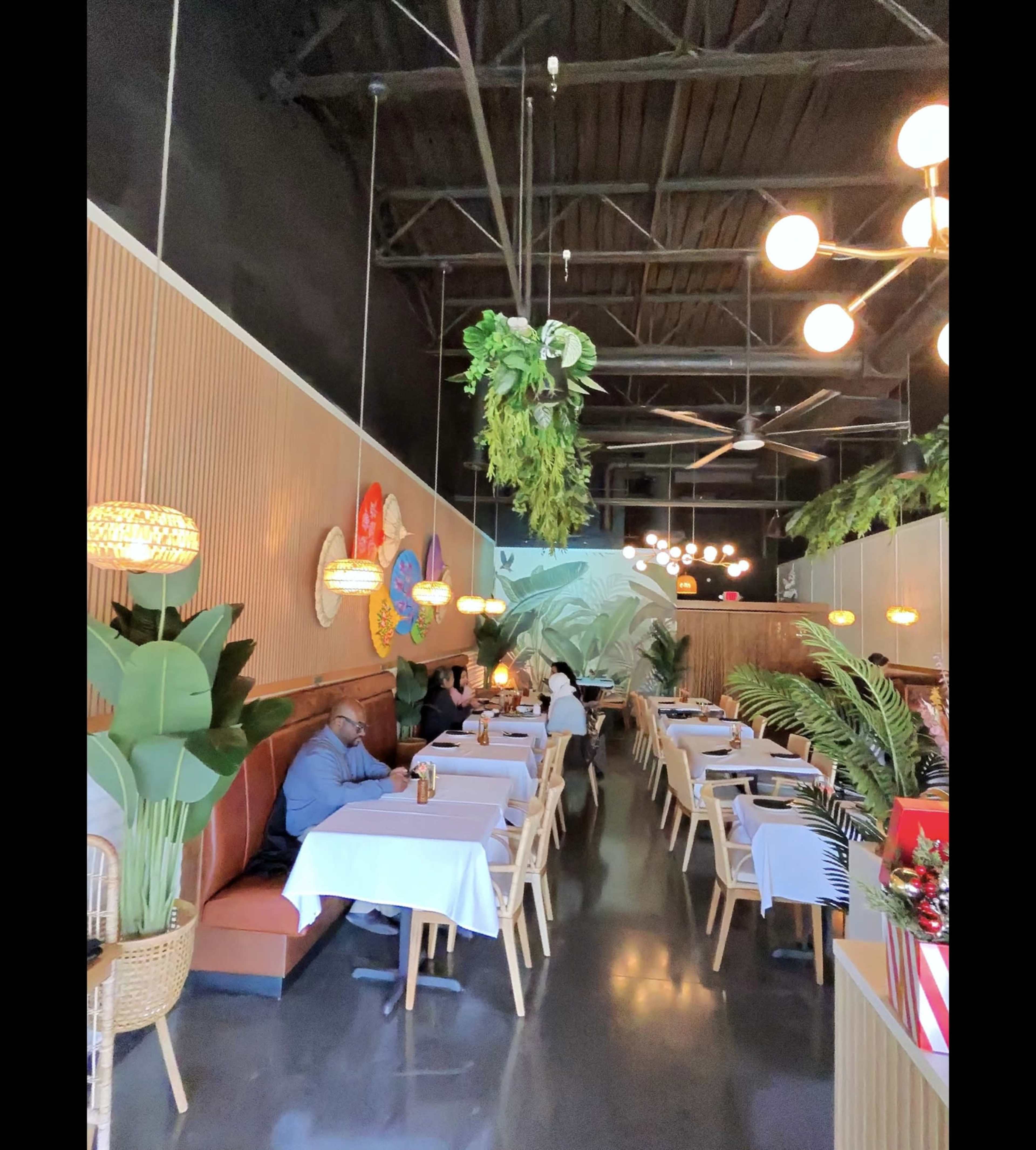 The image shows a modern restaurant interior with tables set for dining, featuring greenery and decorative lighting along the walls.